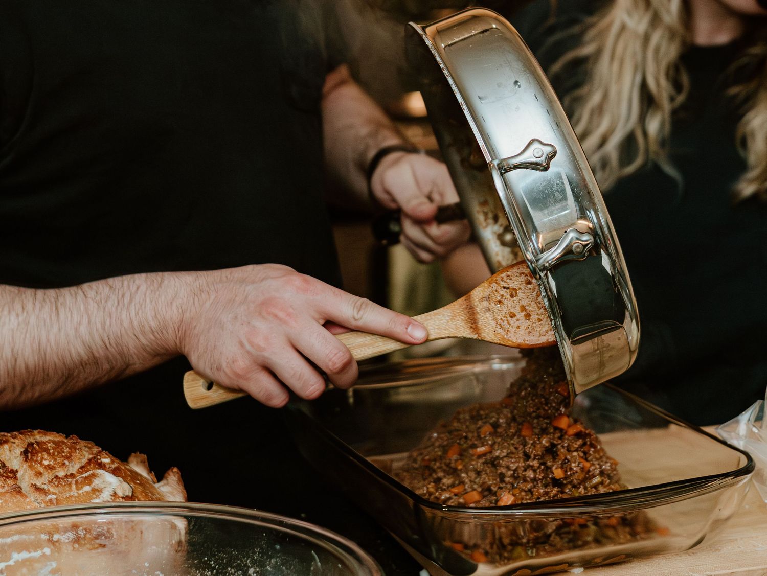 Someone pours a hot liquid from a silver pitcher onto a baking sheet filled with chopped nuts and spices.