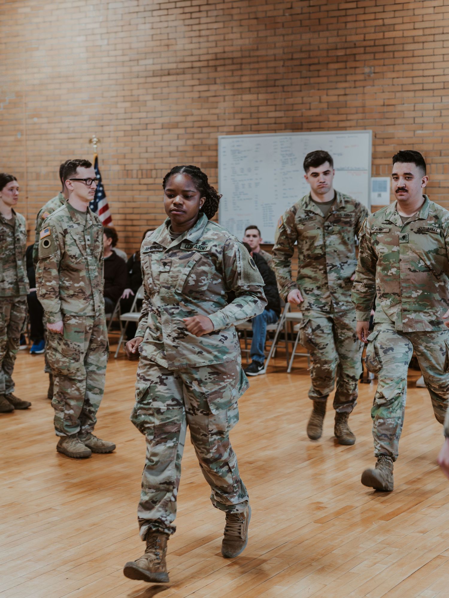 Service members in combat uniforms participate in a training exercise on a hardwood floor in an indoor facility.
