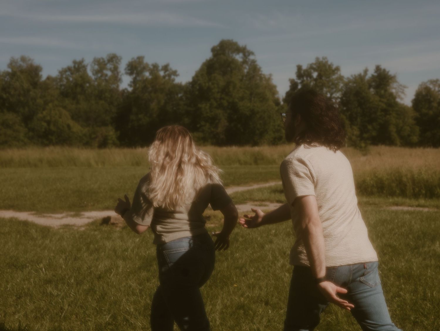 Two people hold hands while walking through a sunny meadow surrounded by trees in a vintage-style photo.