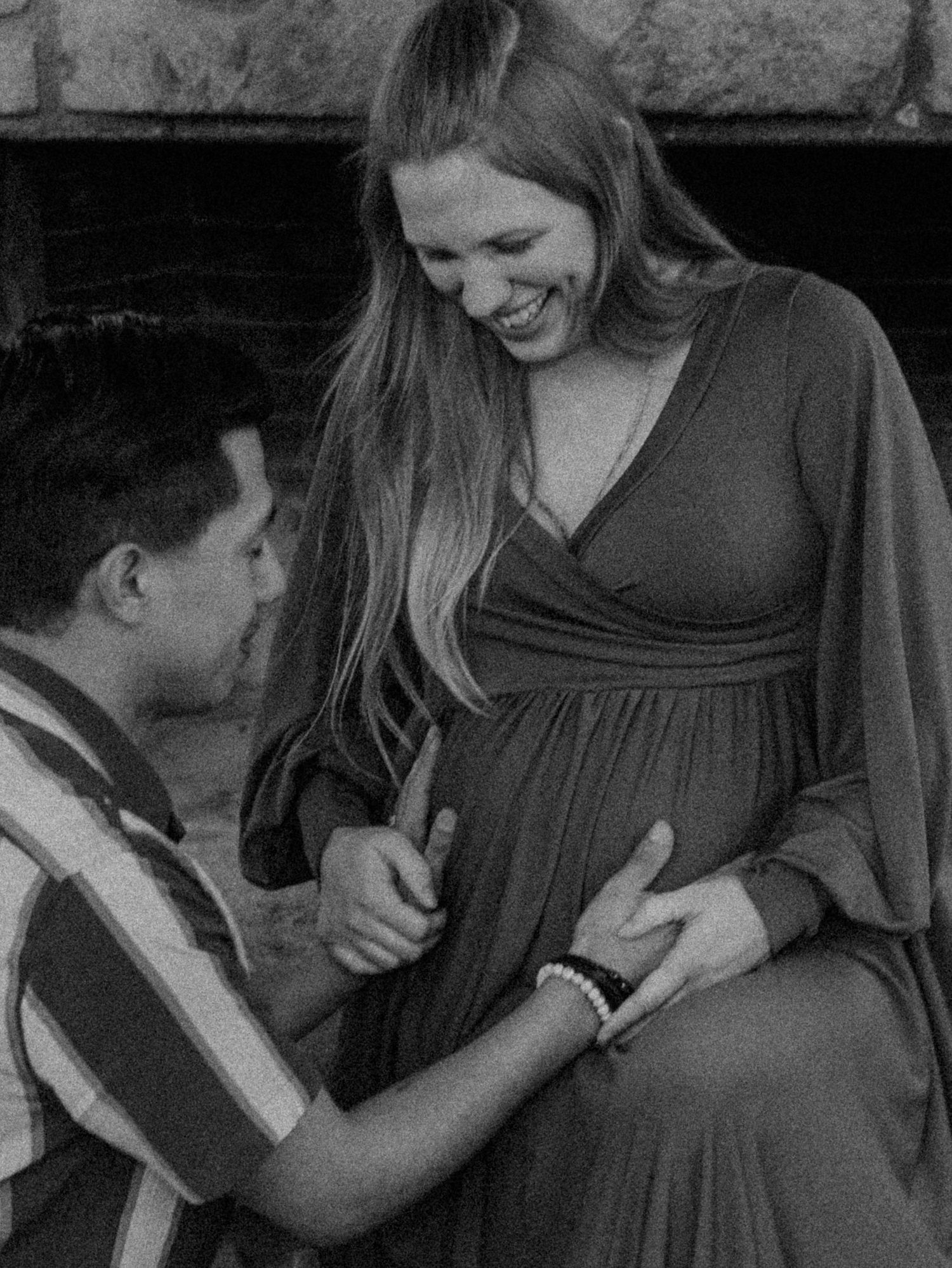 A candid black and white photo showing a tender moment of a young boy touching a pregnant woman's belly as they share laughter.