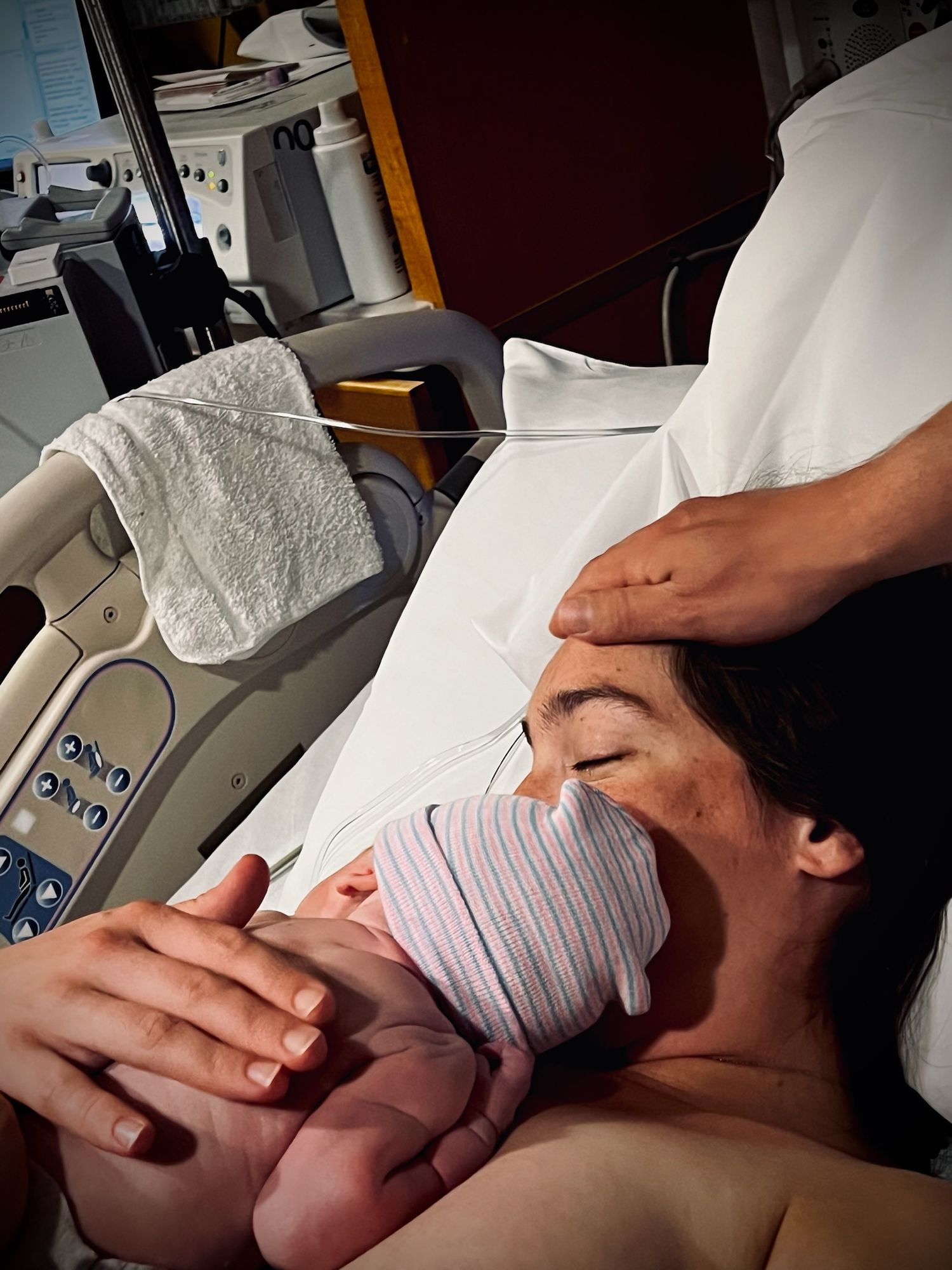 A newborn baby wearing a hospital cap being cradled against skin in a medical setting.