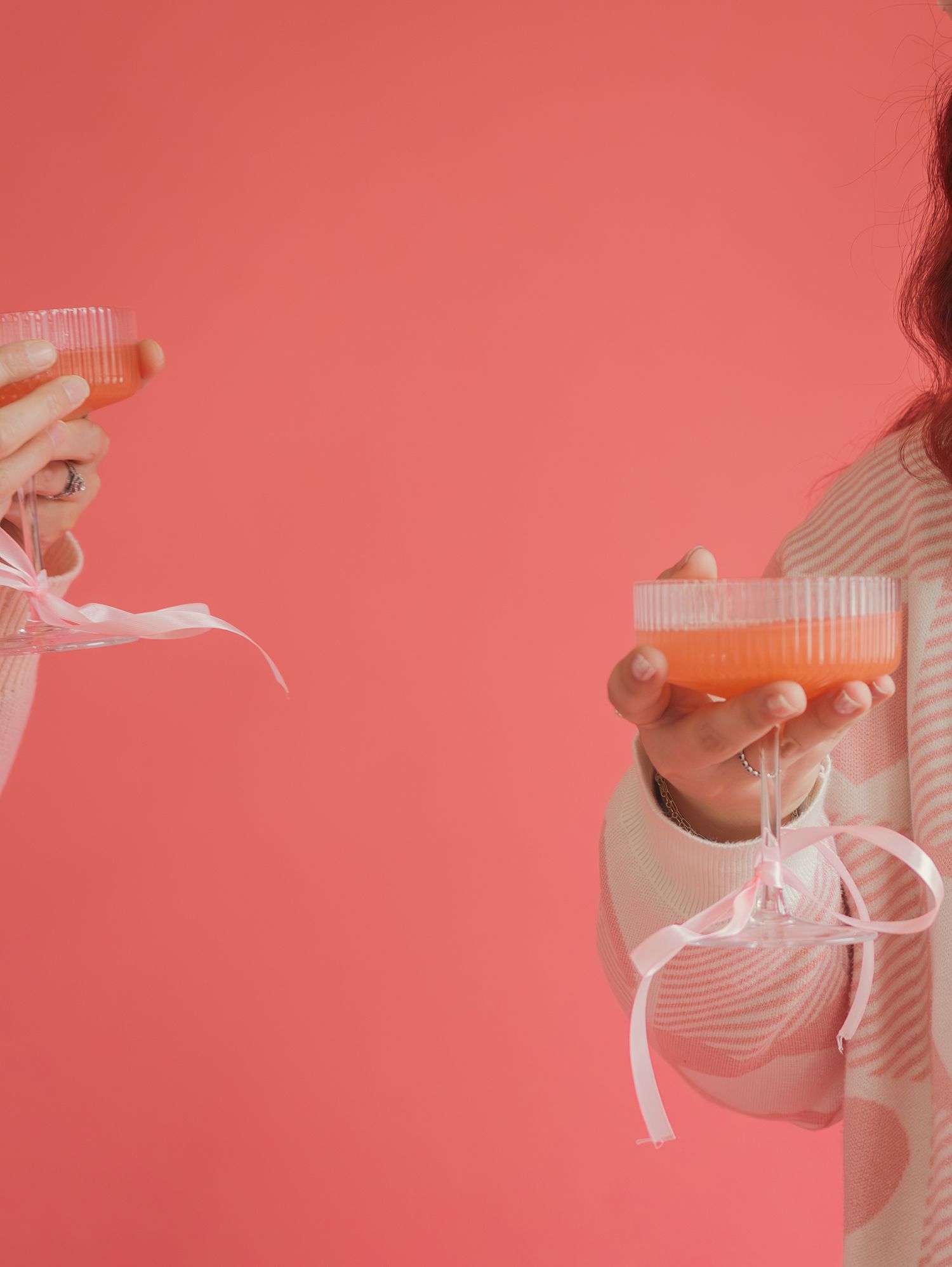 Two people holding pink drinks with ribbons against coral colored background.