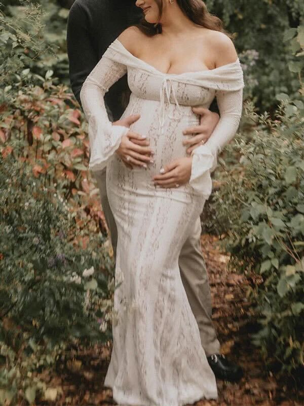 Intimate maternity photoshoot outdoors showing off-shoulder white lace gown against lush greenery background.