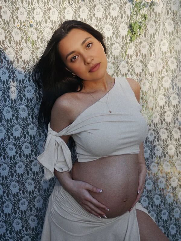 An expectant person in a flowy white wrap dress poses against a textured wall backdrop during a maternity photoshoot.