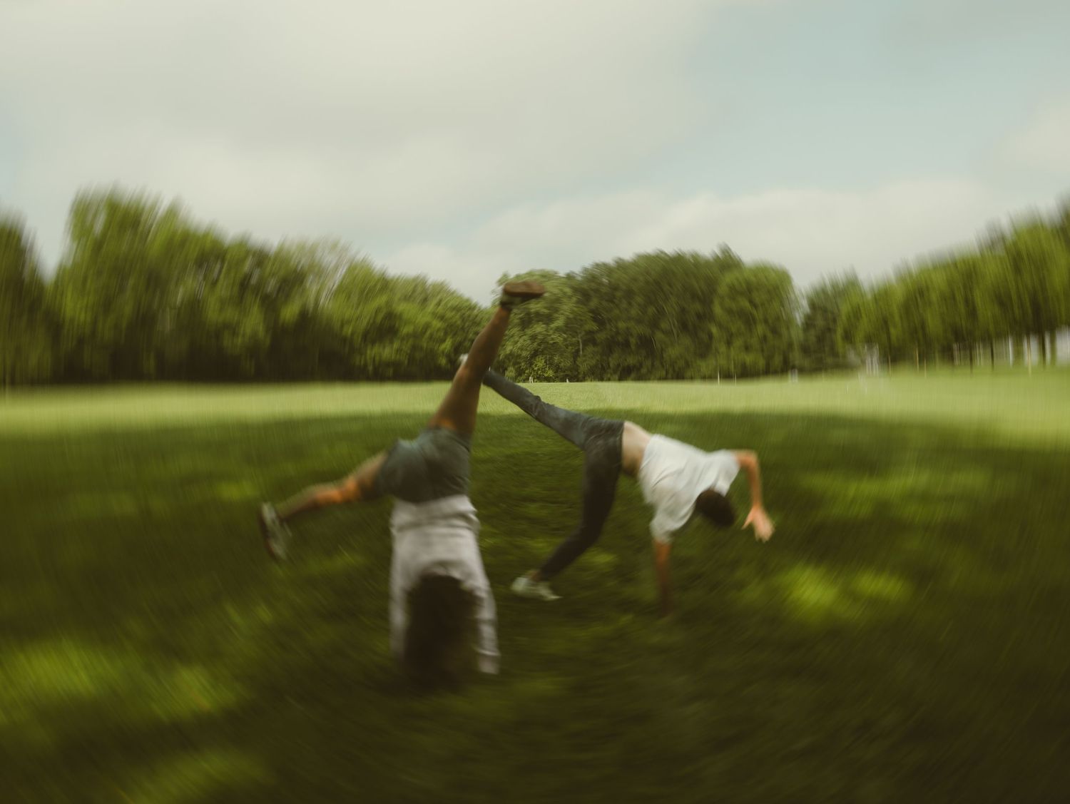 Two children doing cartwheels together on a lush green grass field in a park on a cloudy day.