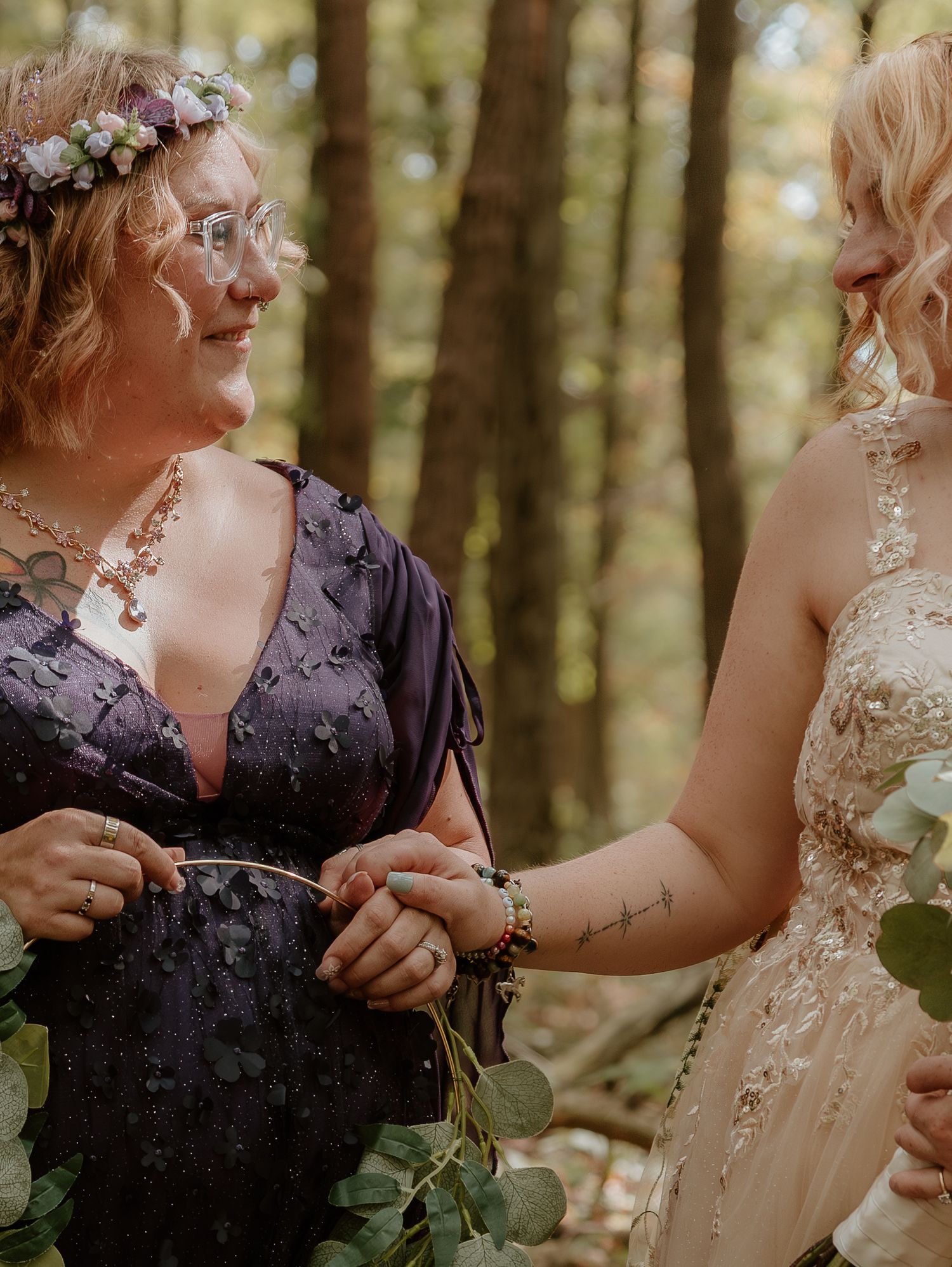 Wedding party members in purple and cream dresses share a moment in a sunlit forest.