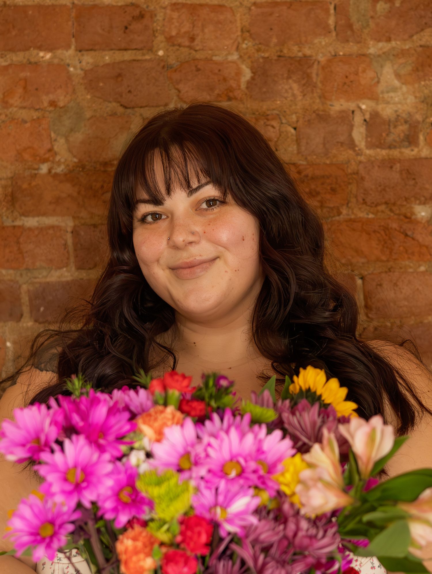 A person smiling next to a vibrant bouquet of colorful flowers against a brick wall.