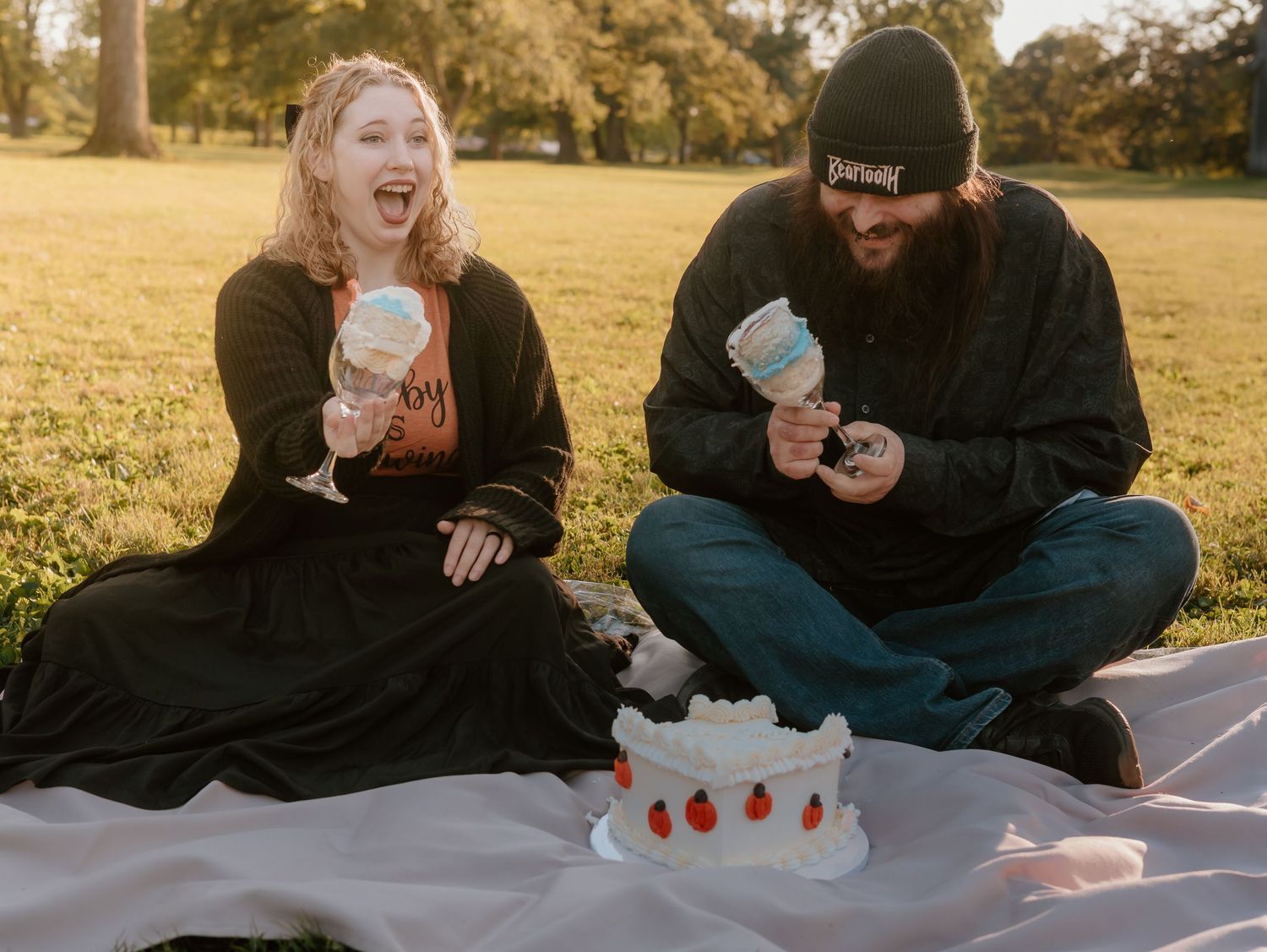 A couple laughs together while sitting on a blanket in a grassy field at sunset.