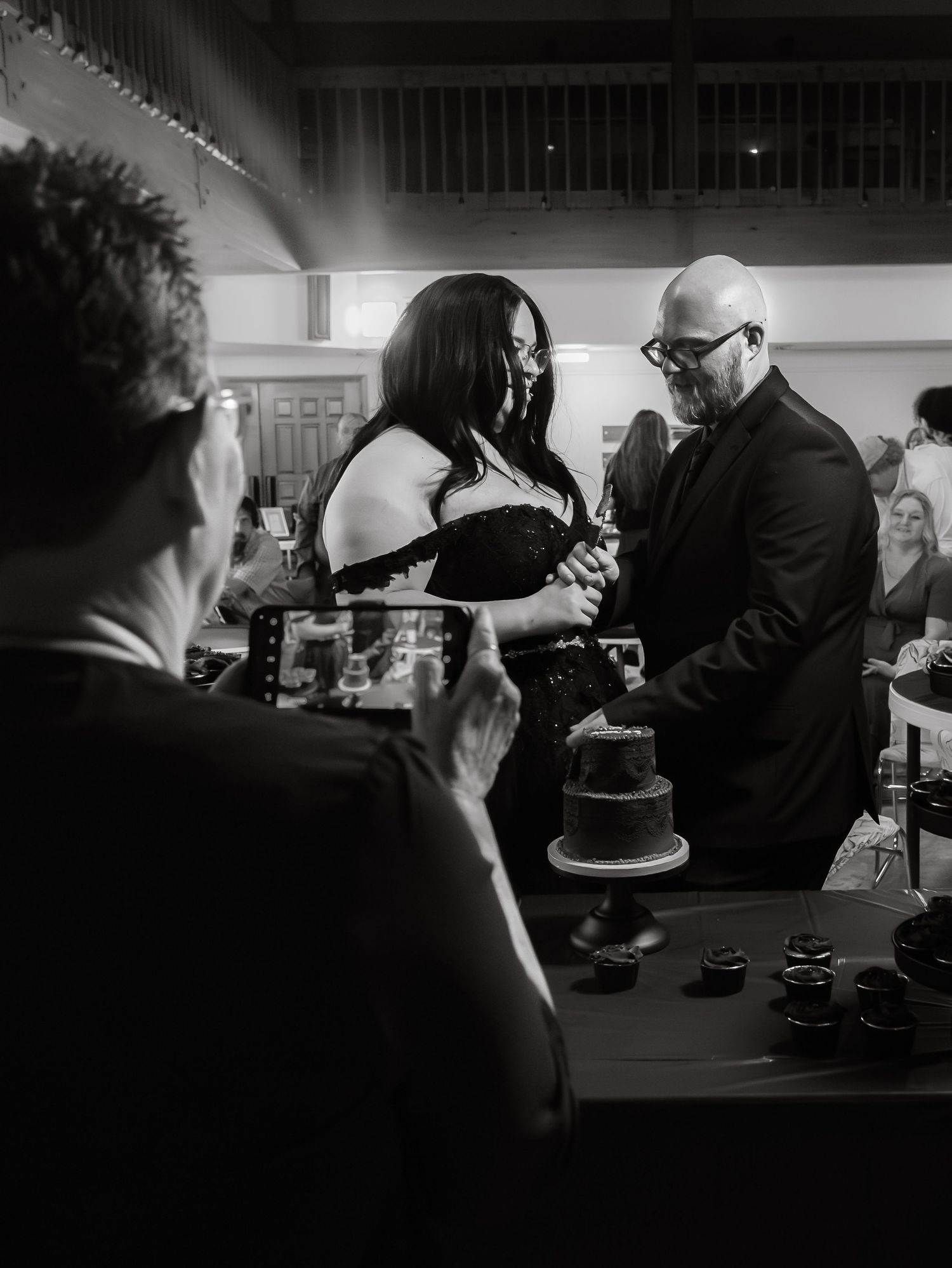 Black and white photo of bride and groom cutting dark wedding cake at gothic reception.