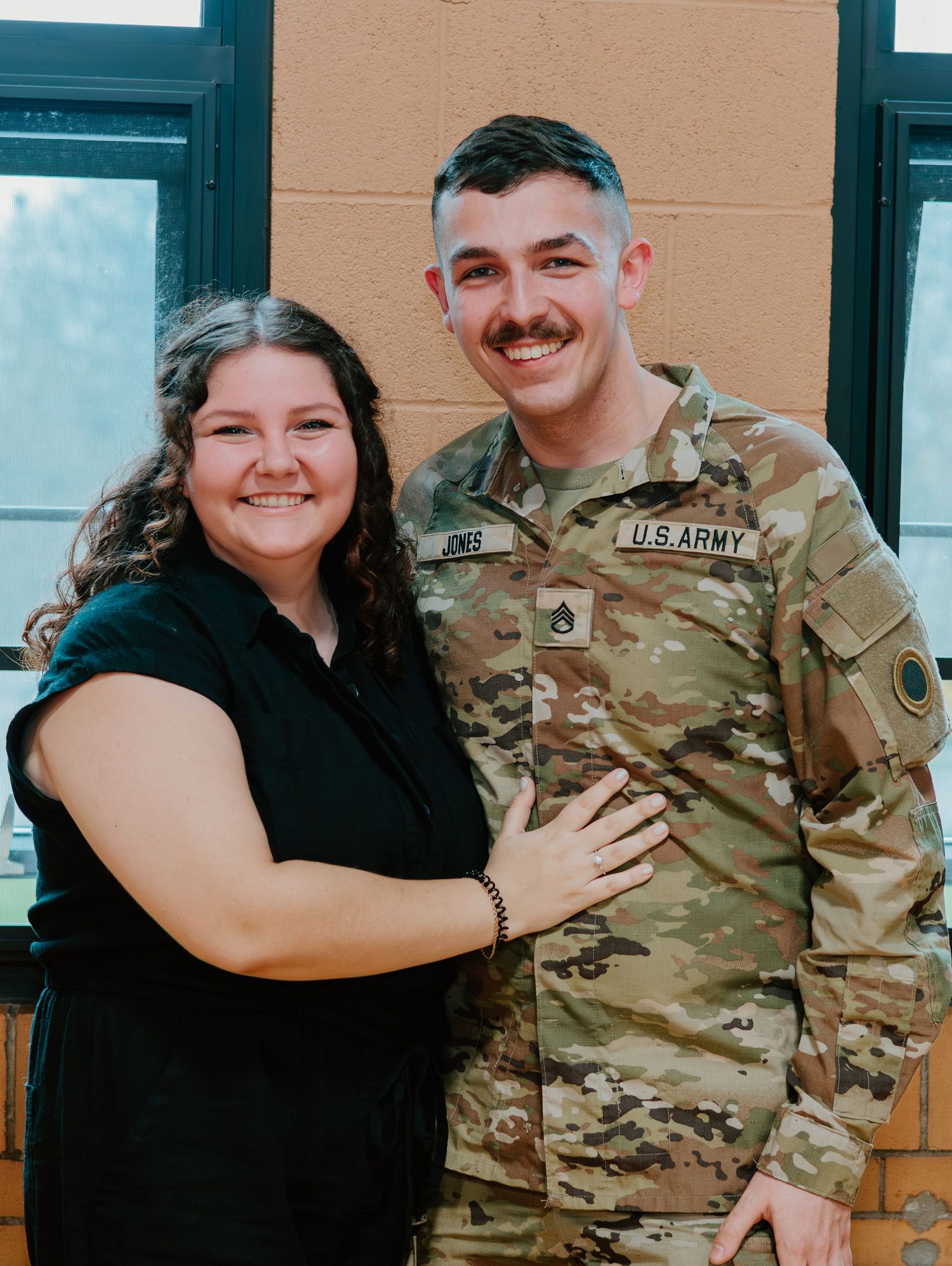A couple poses together, with one wearing a black dress and the other in U.S. Army camouflage uniform.