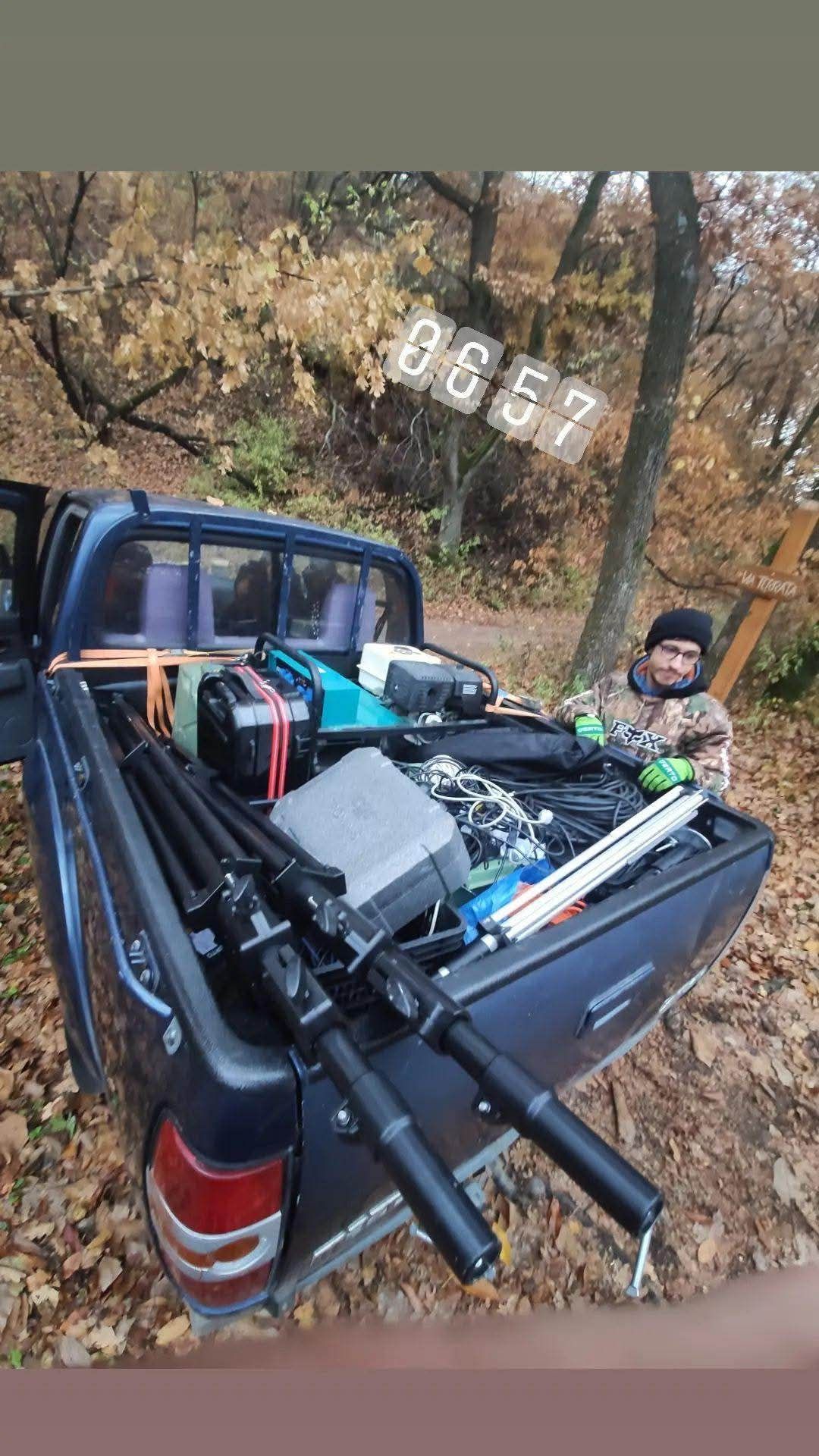 A pickup truck bed loaded with various outdoor equipment including fishing rods, gear, and tools on a wooded trail.