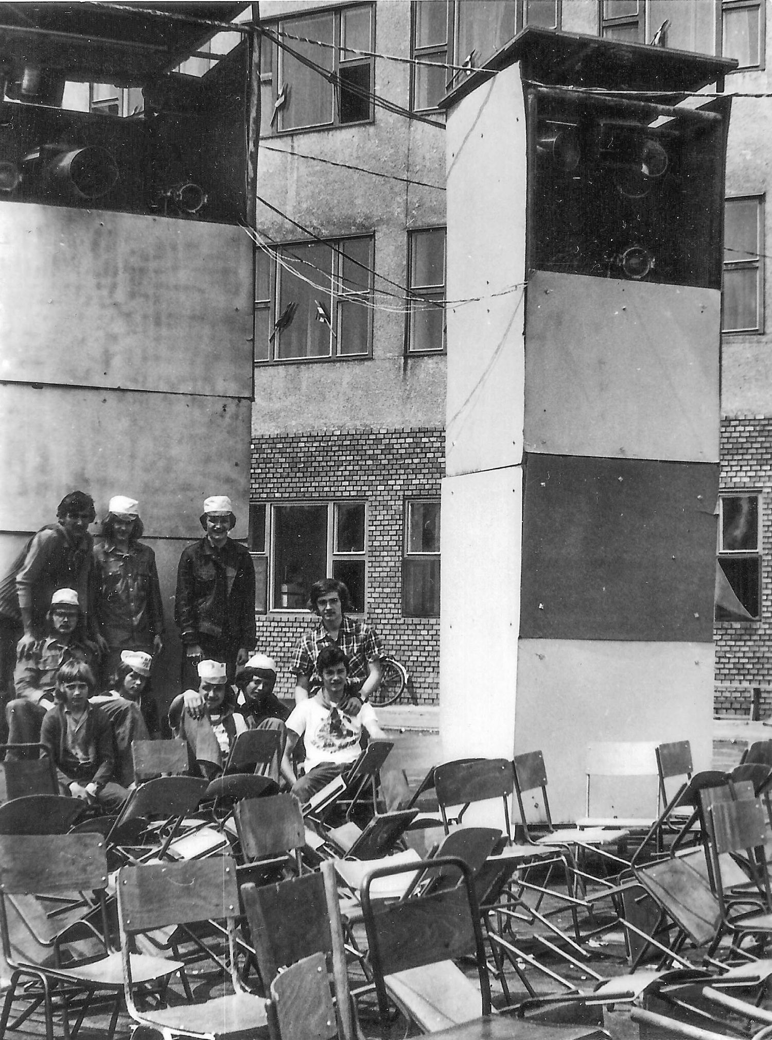 Monochrome photograph shows people gathered around chairs and a striped wall at a multi-story apartment building.