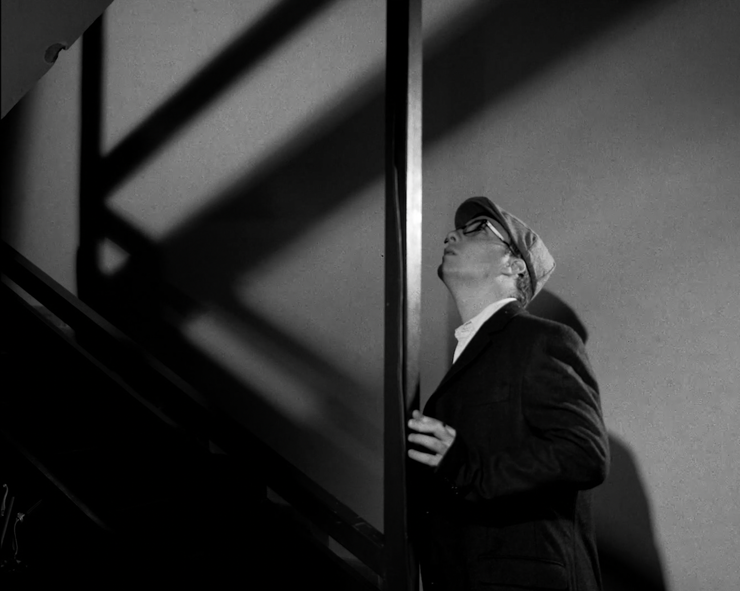 Dramatic black and white photograph showing a figure looking up while standing in a stairway with stark shadows.
