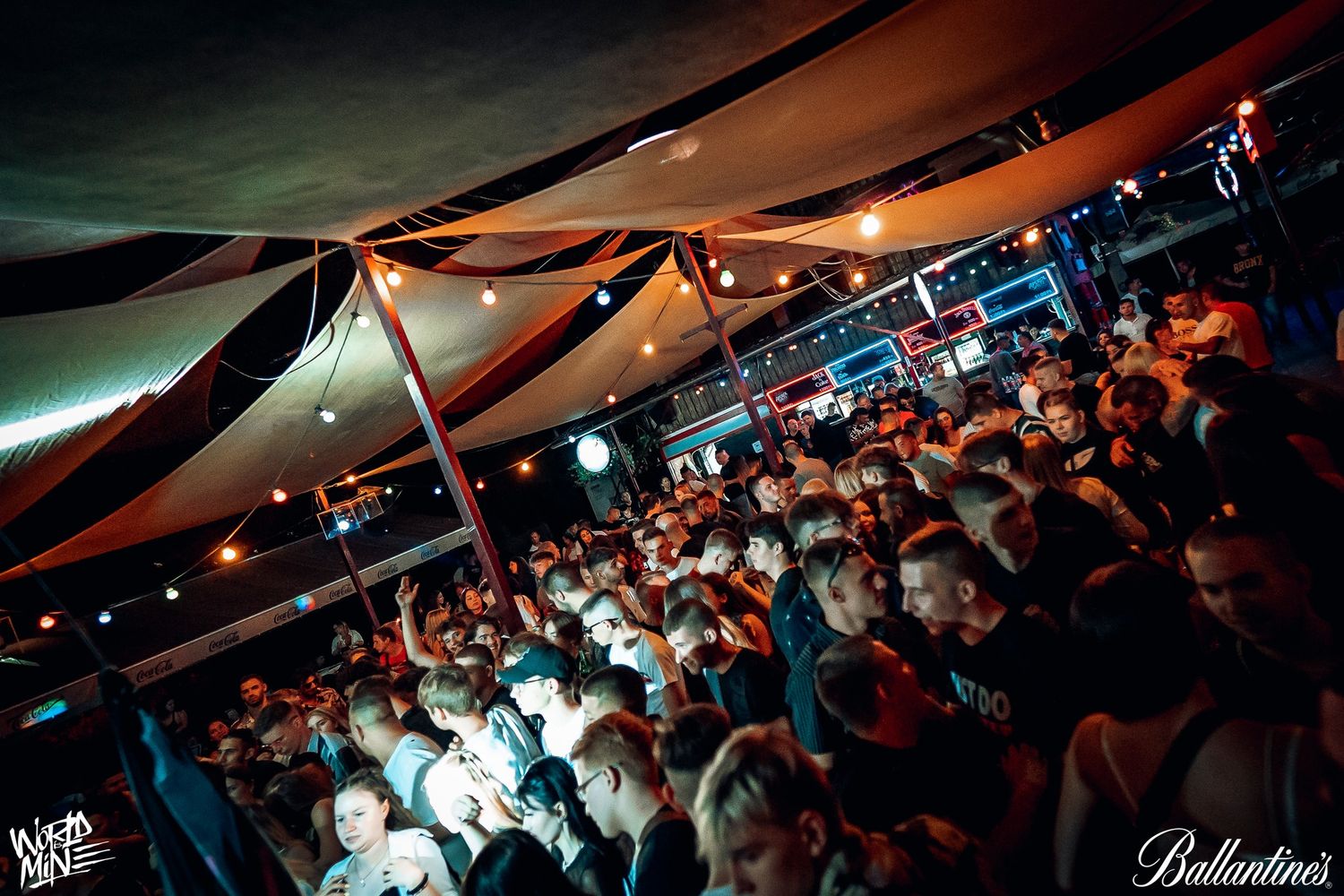A crowded nightclub dance floor under orange tent canopies with string lights and blue lighting illuminating the crowd.