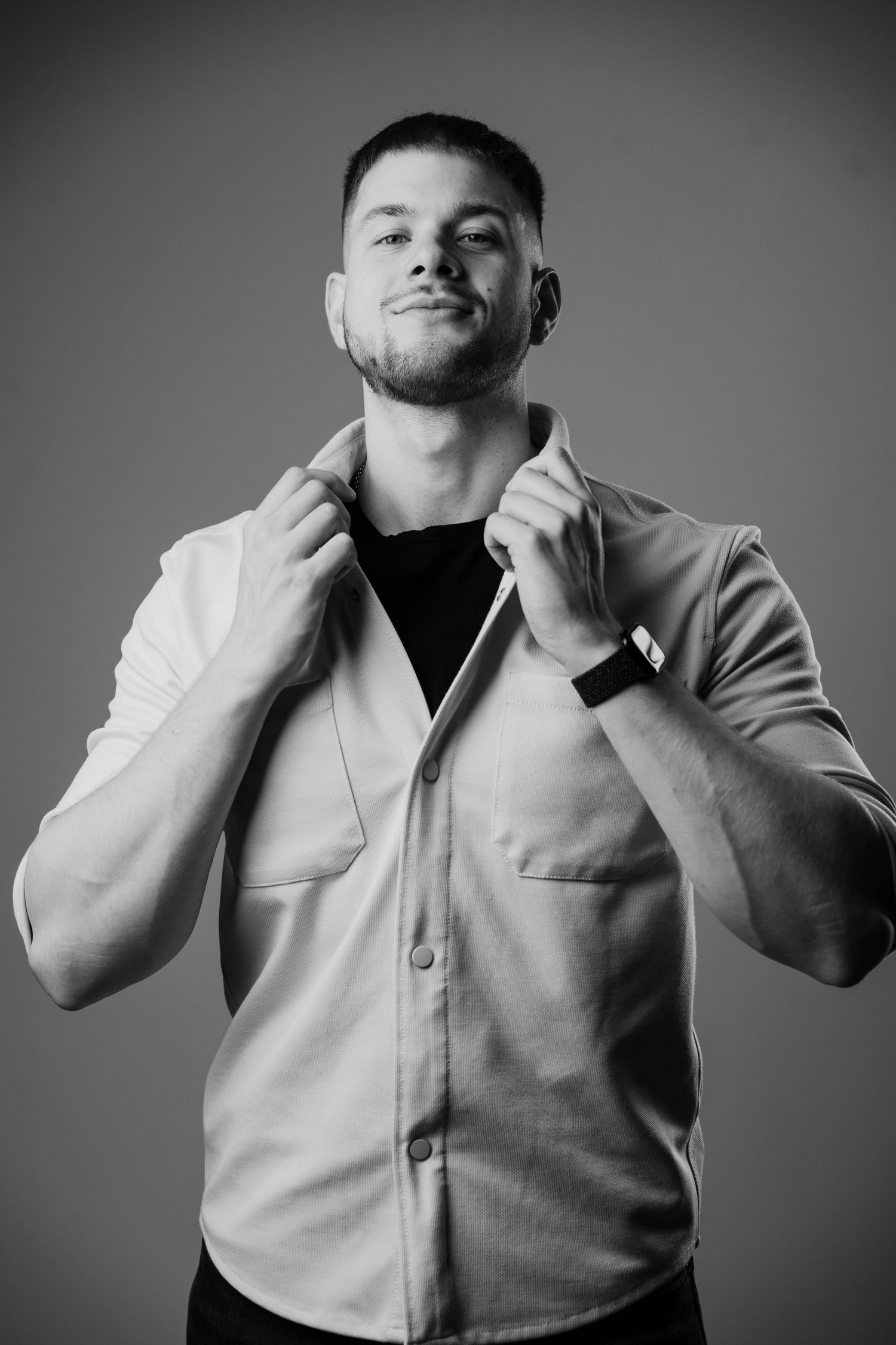 Black and white portrait of someone adjusting their collar while wearing a light button-up shirt and dark undershirt.