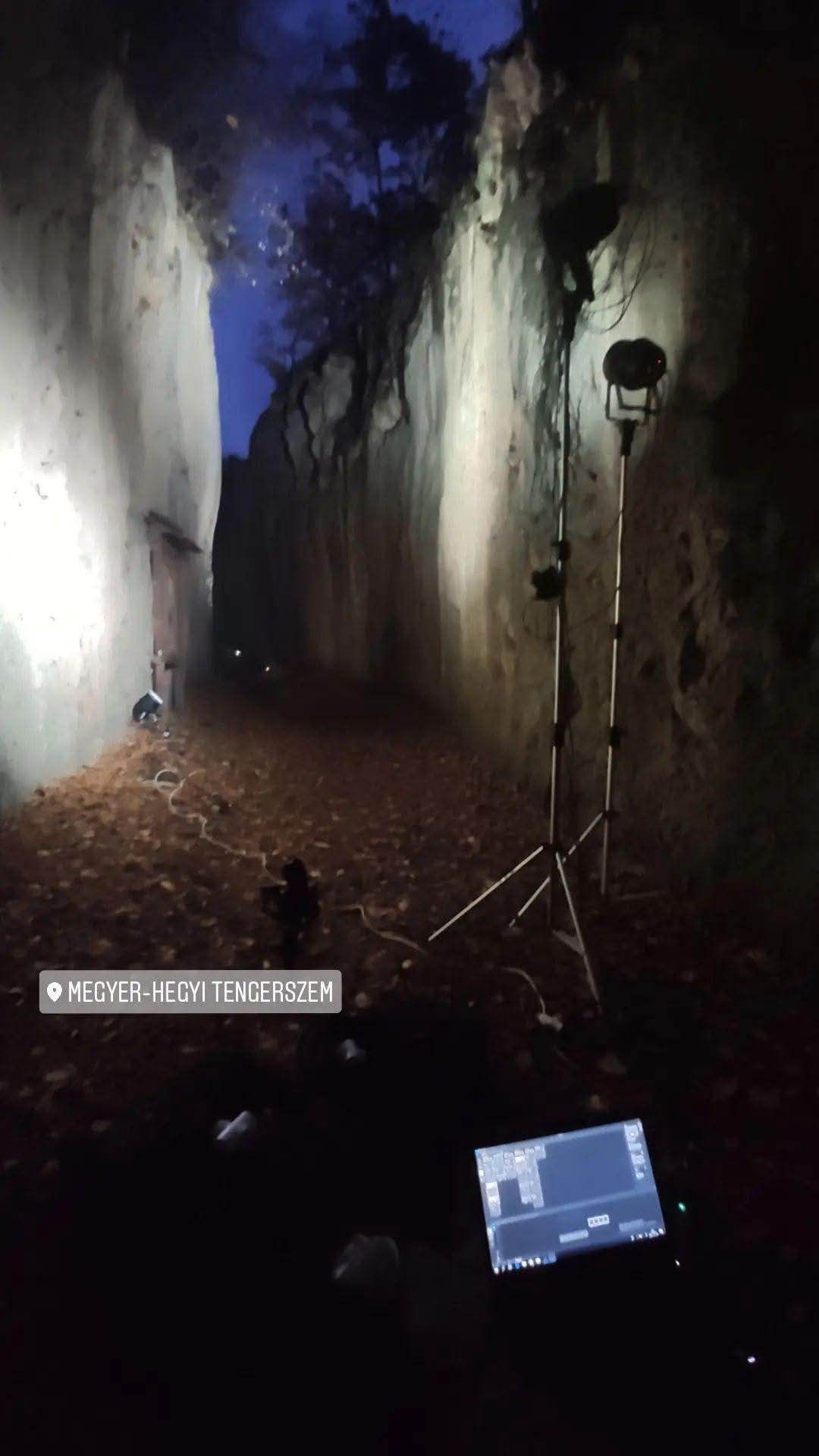 Behind-the-scenes lighting setup for a film or photo shoot in a dark cave with tall rock walls and spotlights mounted on stands.