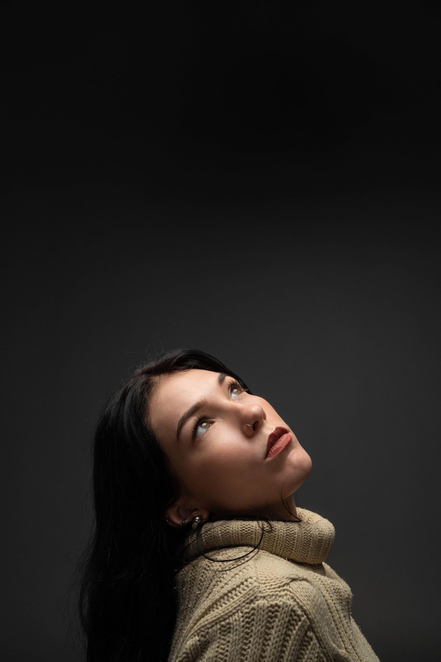 Dark artistic portrait of someone in beige sweater looking upward against gray background.
