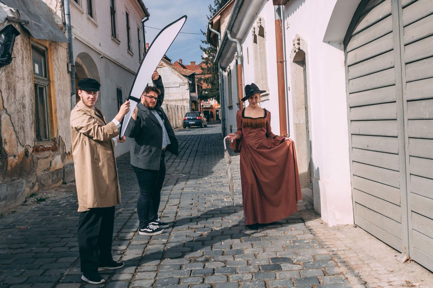 Three people in vintage historical costumes pose on a cobblestone street in an old town, with a giant scythe prop.