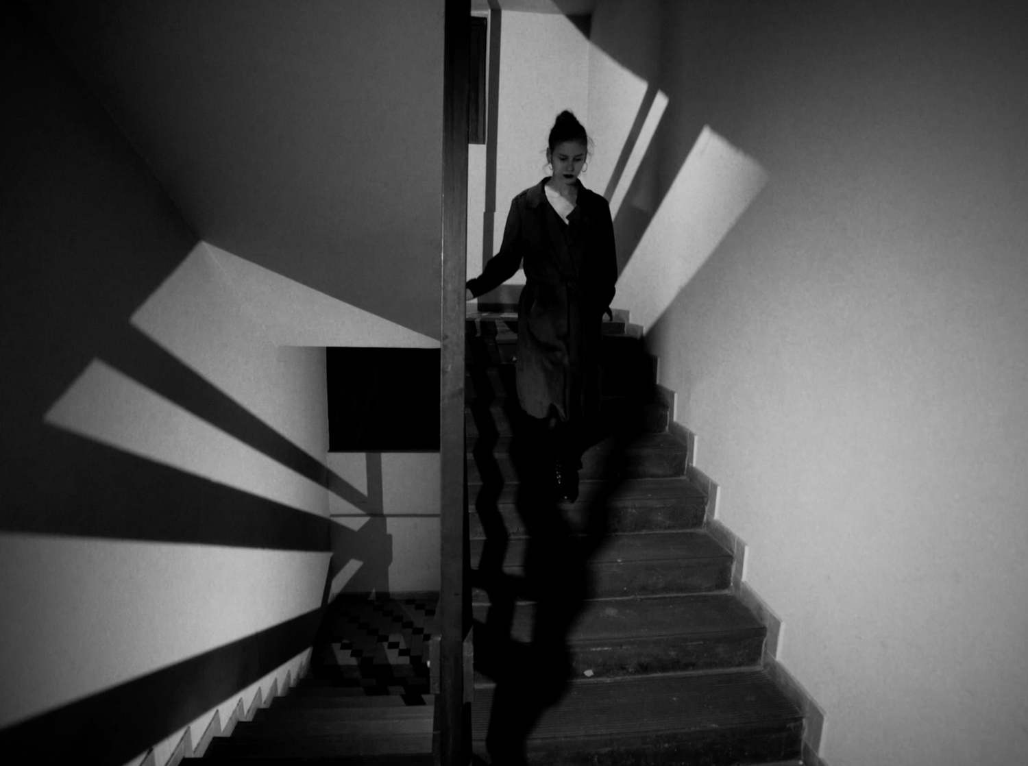 A dramatic black and white shot of a silhouetted figure ascending a staircase with angular shadows along the wall.