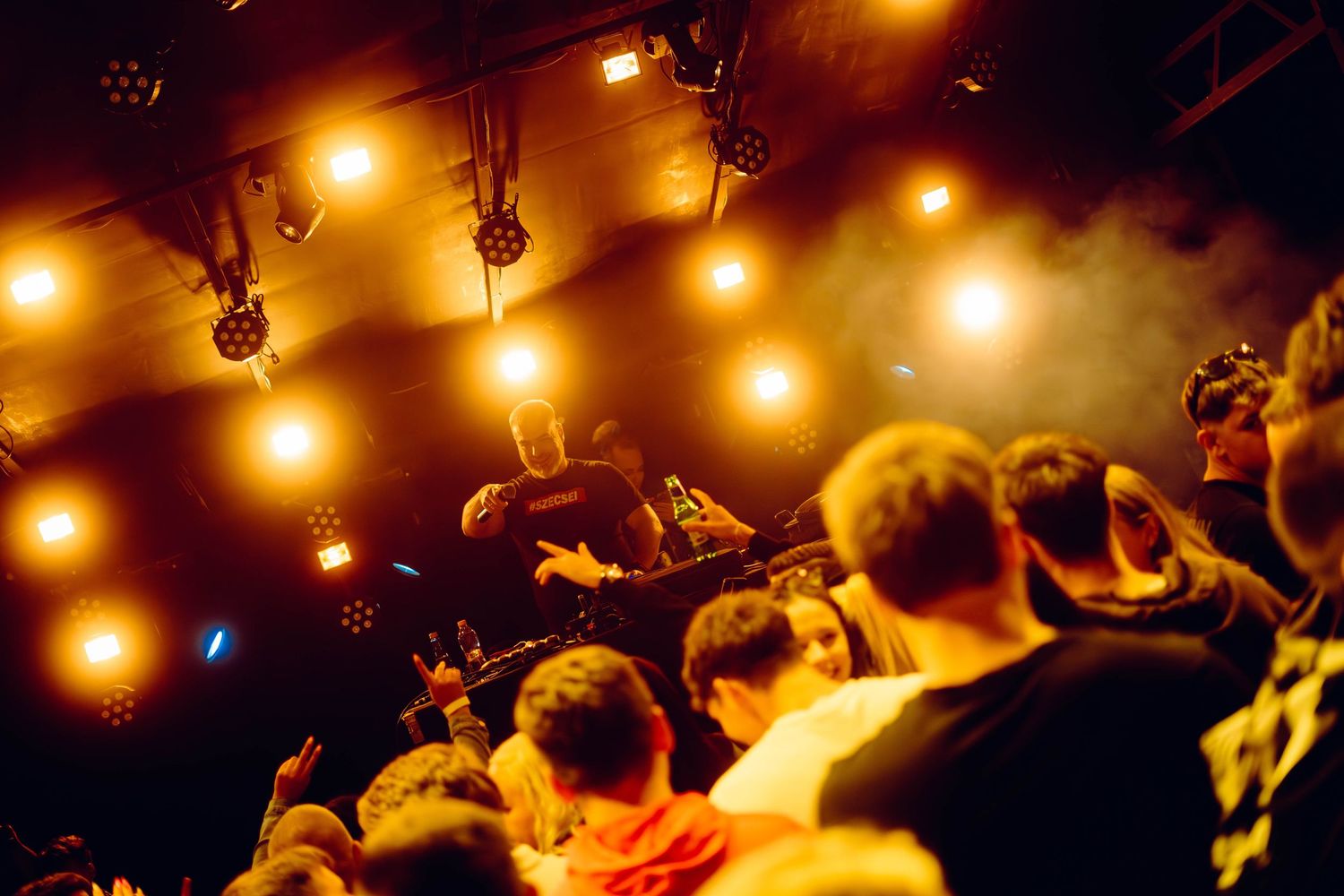Concert crowd silhouettes illuminated by warm overhead stage lights in a dark venue during performance.