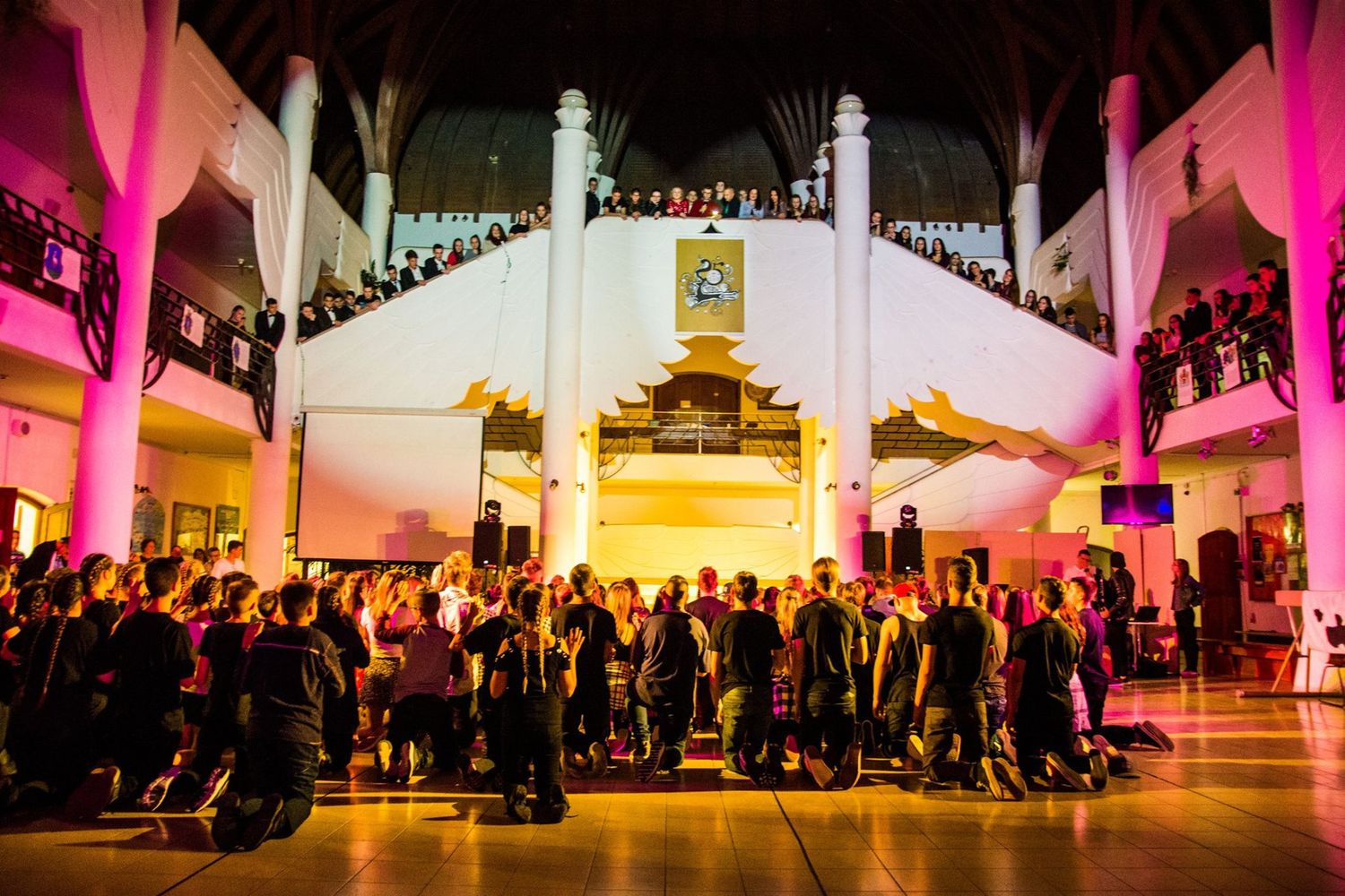 Audience sits on the floor in a dimly lit event space with dramatic pink lighting and high white columns.
