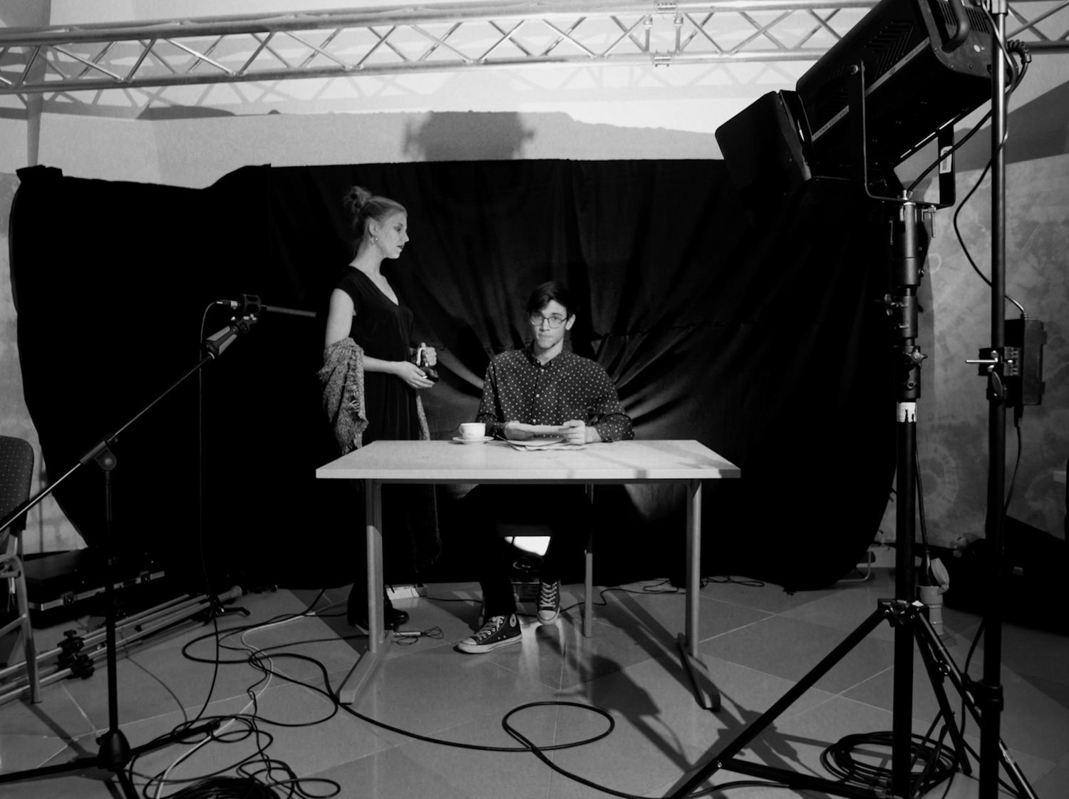 Black and white photo of two people at a table in a studio setting with lighting equipment and dark backdrop.