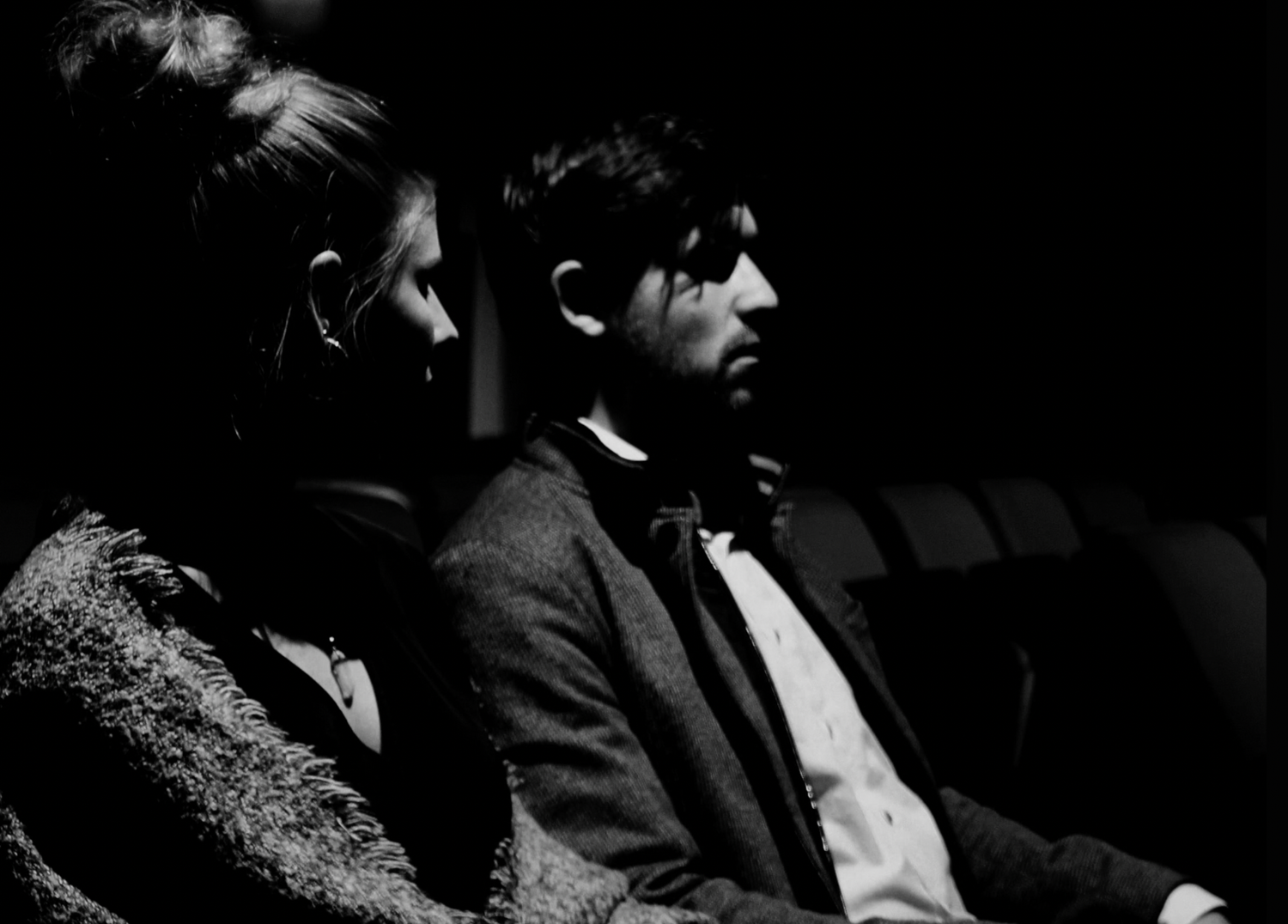 Two people sit together in formal attire in a dark theater or cinema setting, captured in black and white.
