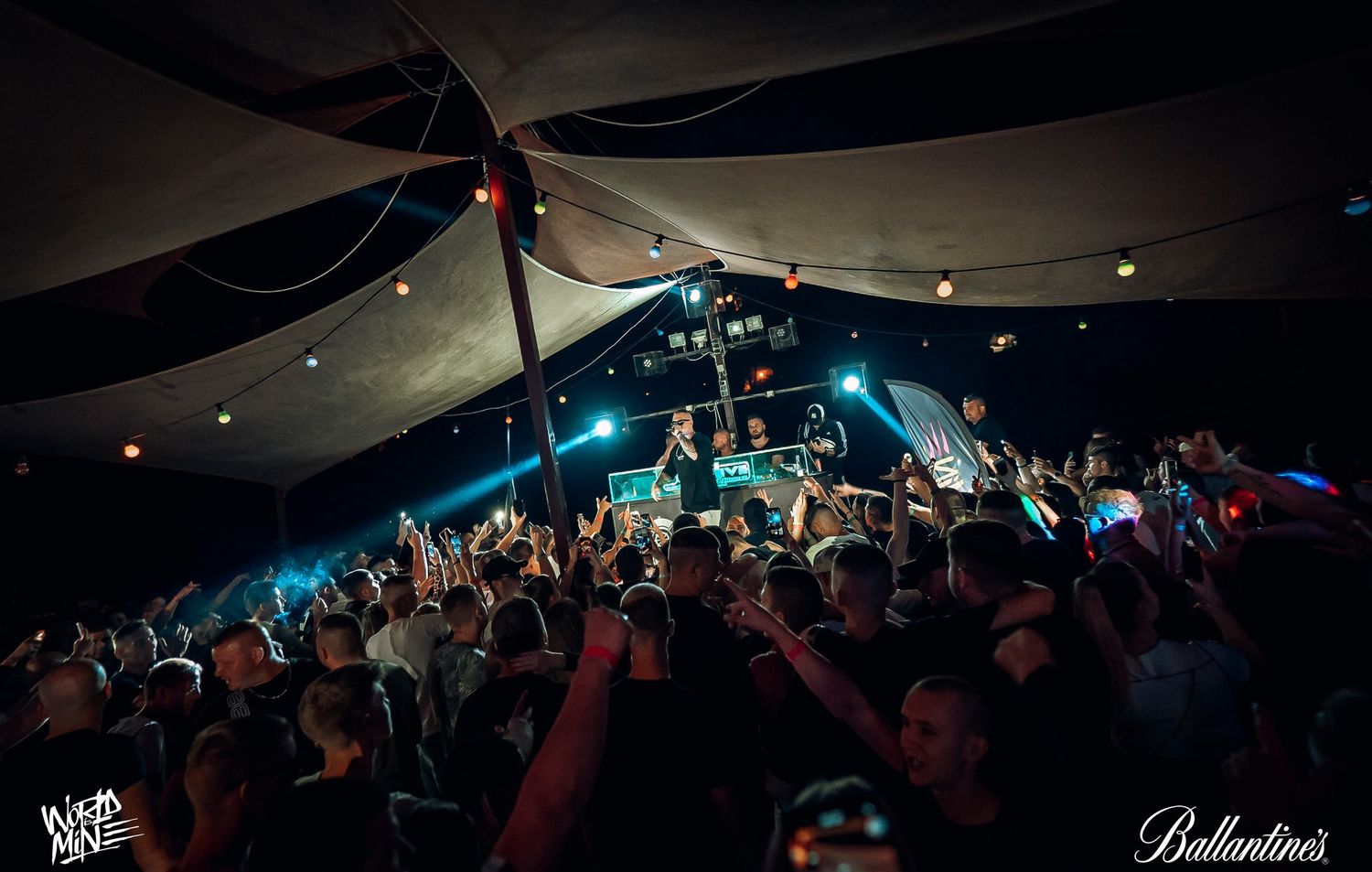 Energetic concert crowd enjoying live music performance under a tent with blue stage lighting and raised hands.