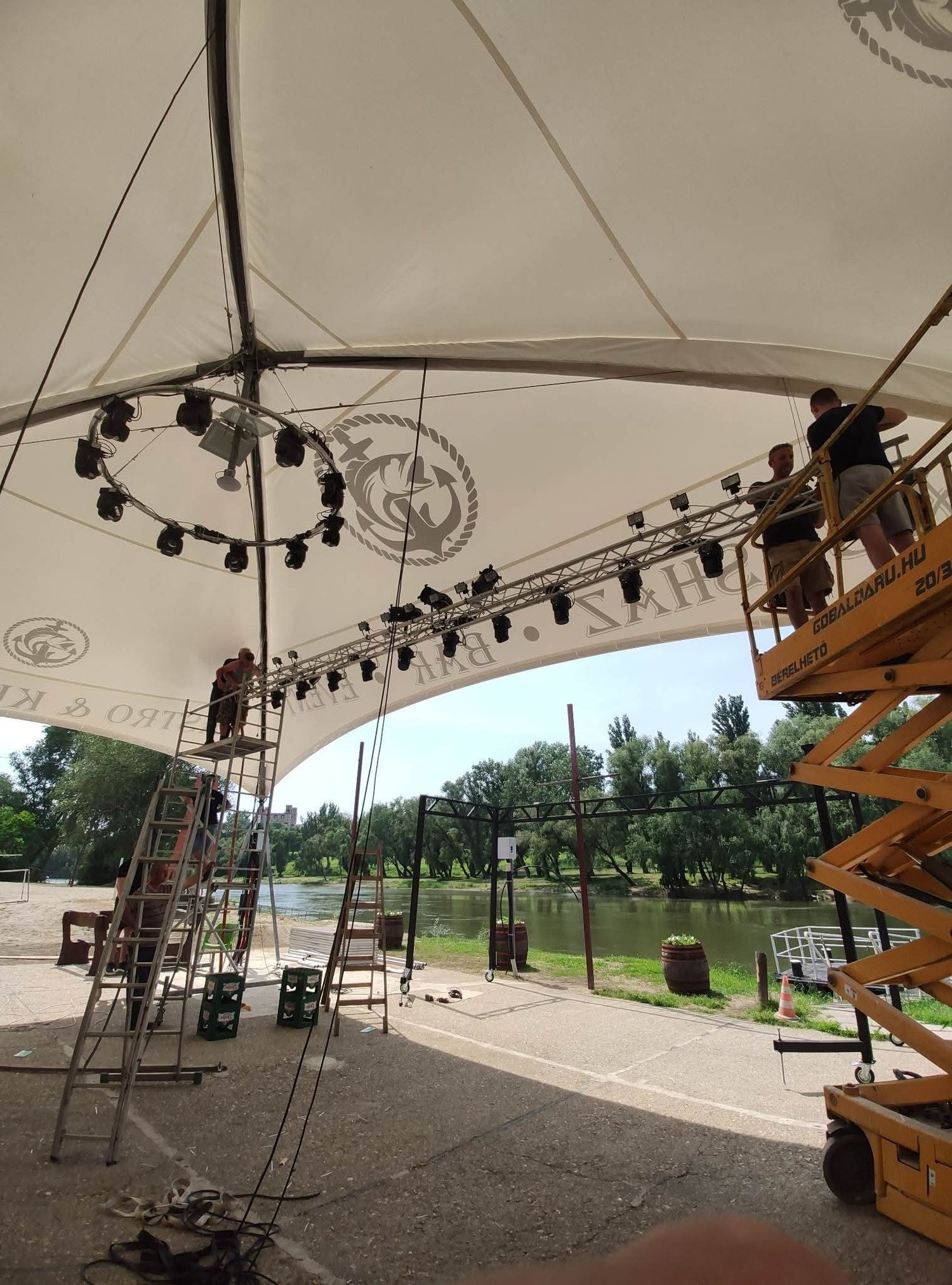 Stage lighting equipment and rigging being set up under an outdoor tent canopy with a scenic park view in the background.