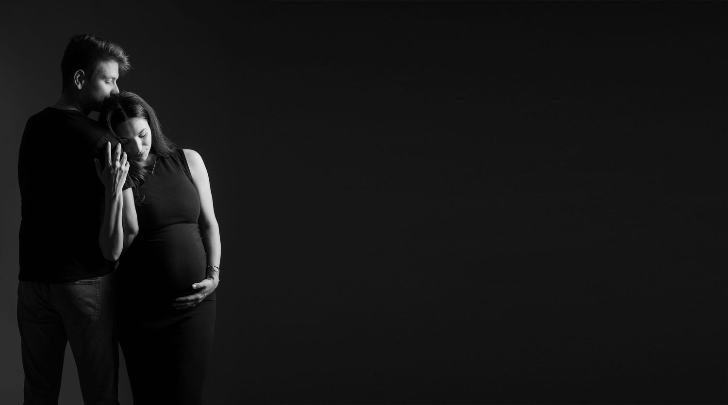 Black and white artistic maternity photo with couple embracing against dark background.