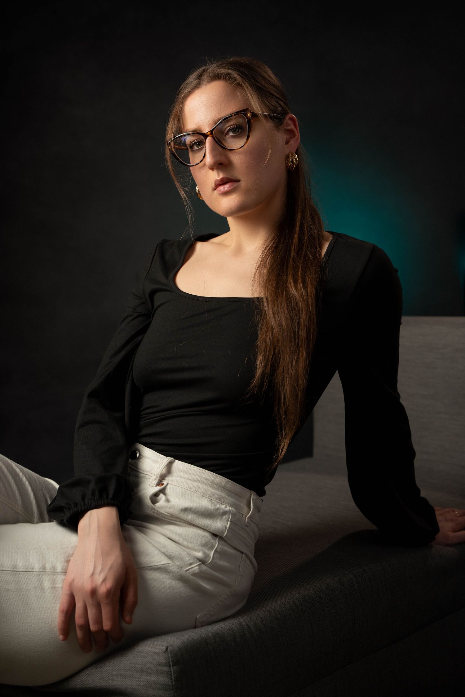 A studio portrait in moody lighting showing a seated figure in glasses, black top and light pants against a dark background.