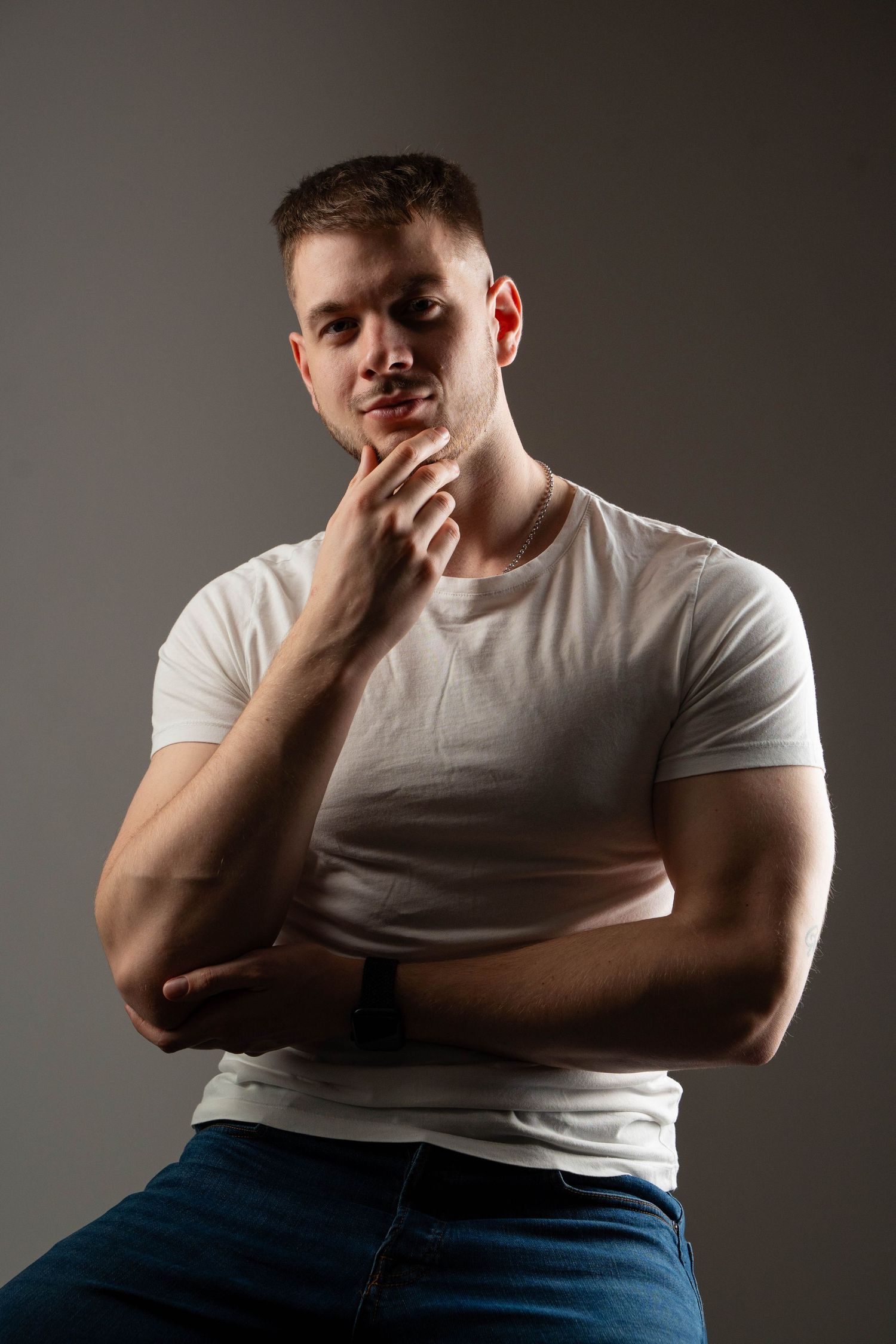 A person in a white t-shirt and jeans poses thoughtfully with hand on chin against a dark background.
