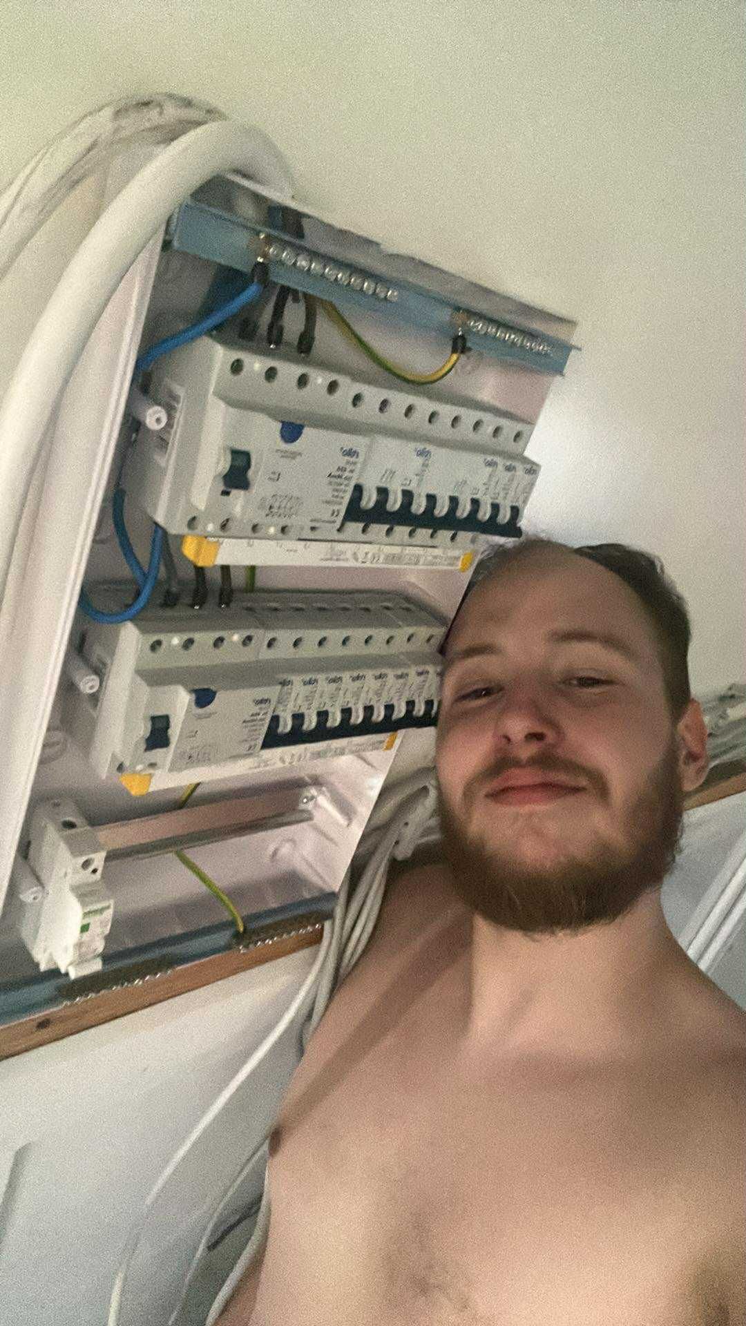 A selfie taken beside an electrical circuit breaker panel showing exposed wiring and circuit switches.