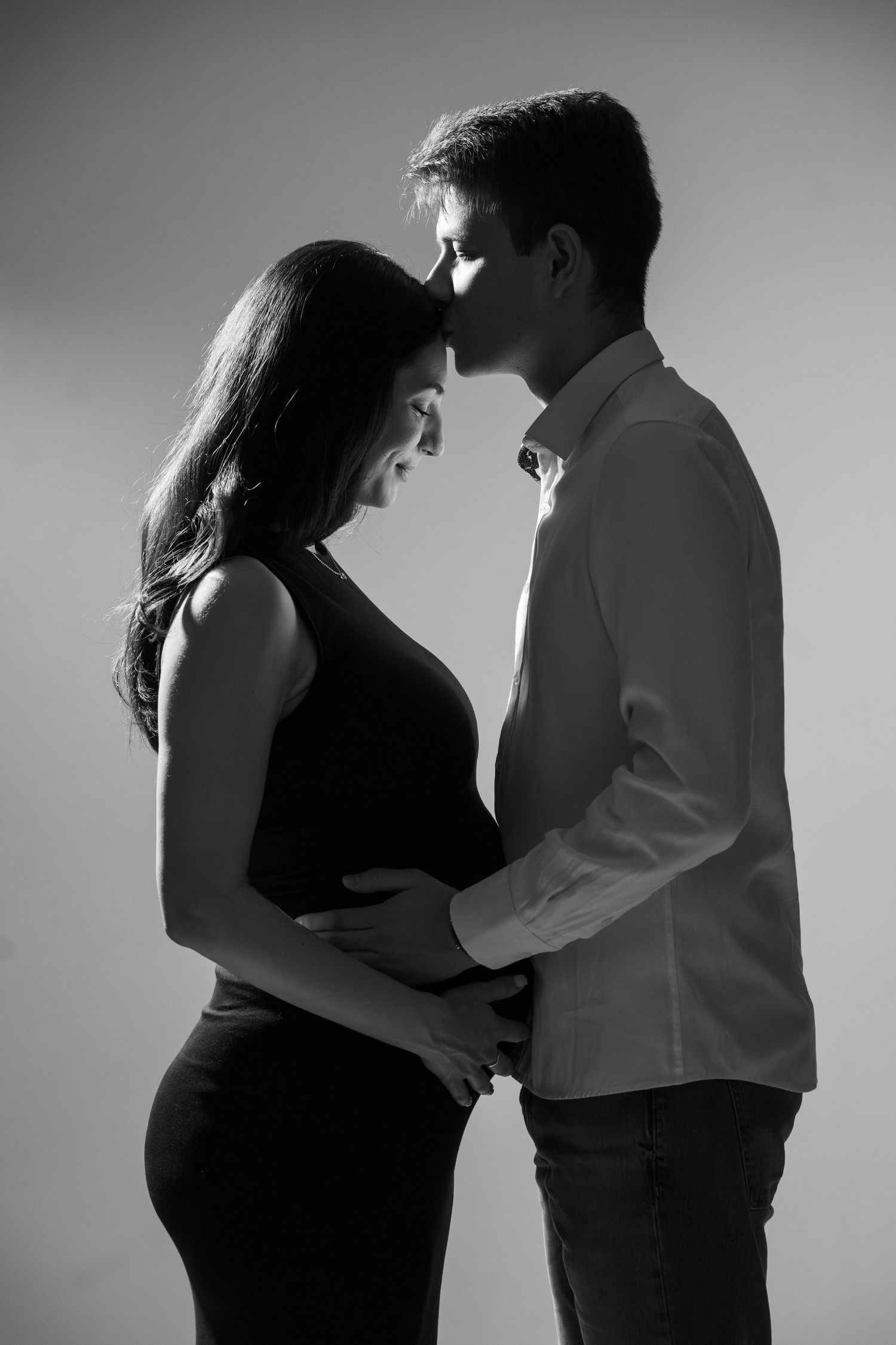 Beautiful black and white maternity photo of couple in intimate embrace against neutral backdrop.
