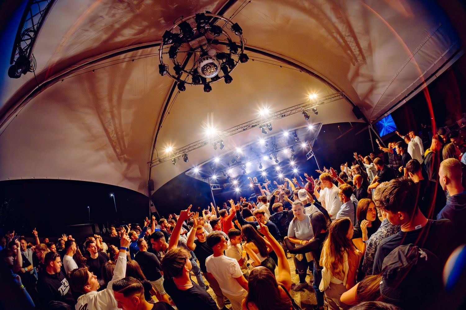Concert crowd with arms raised enjoys live music performance under illuminated venue ceiling.