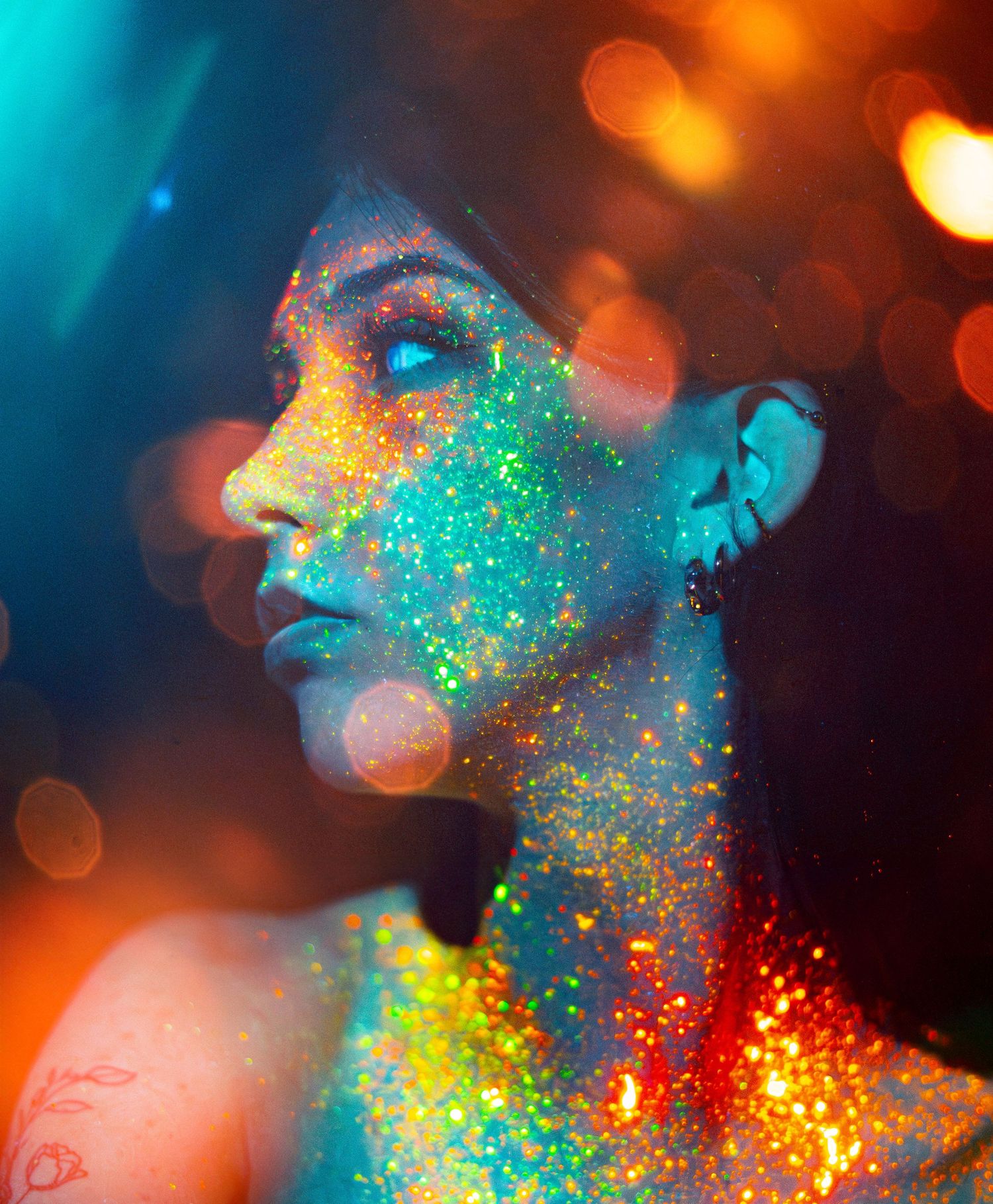 Profile view of someone's face covered in colorful glitter makeup with bokeh lights and turquoise background effects.