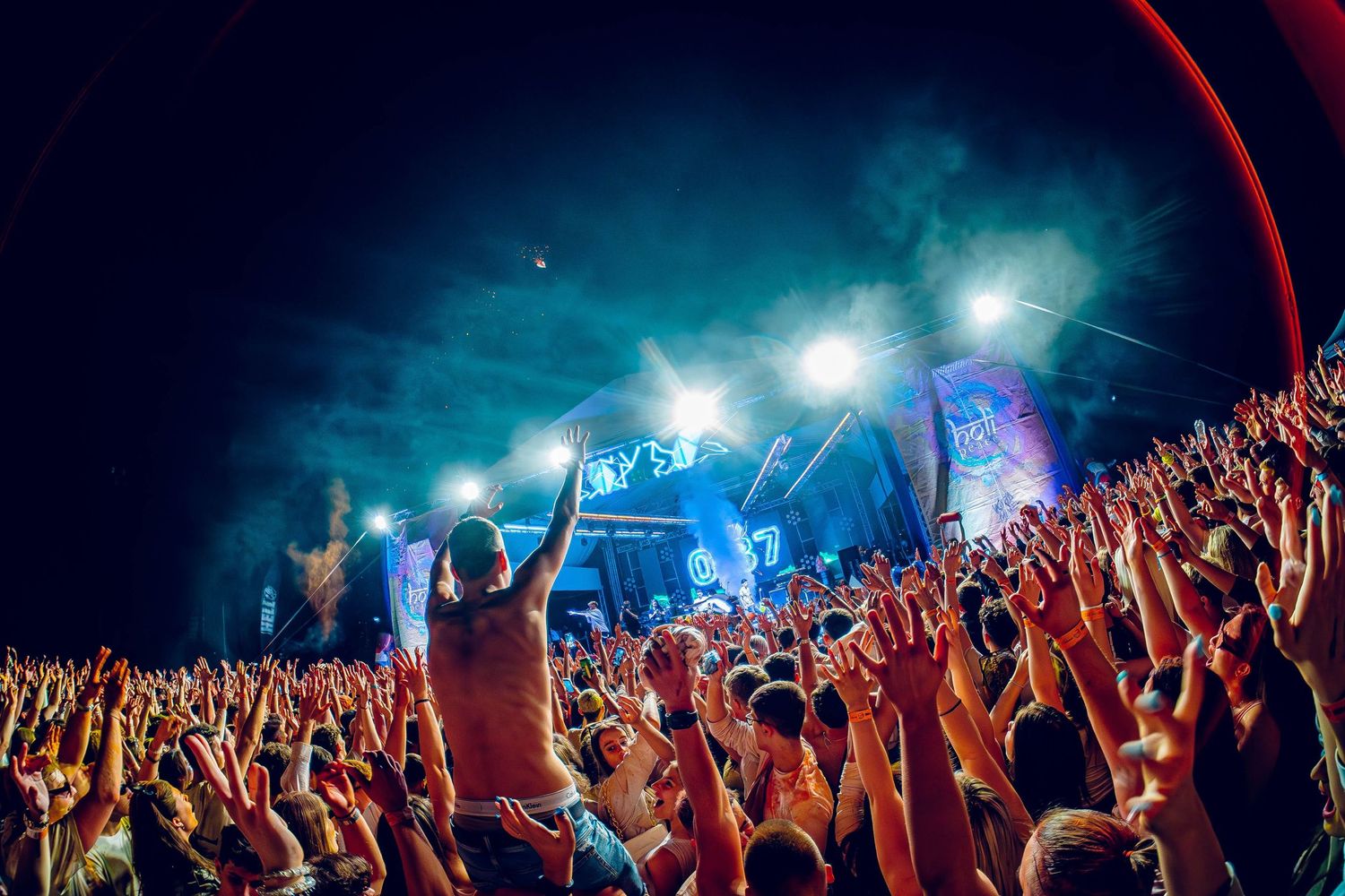 Concert crowd with raised hands silhouetted against blue stage lighting and smoke effects.