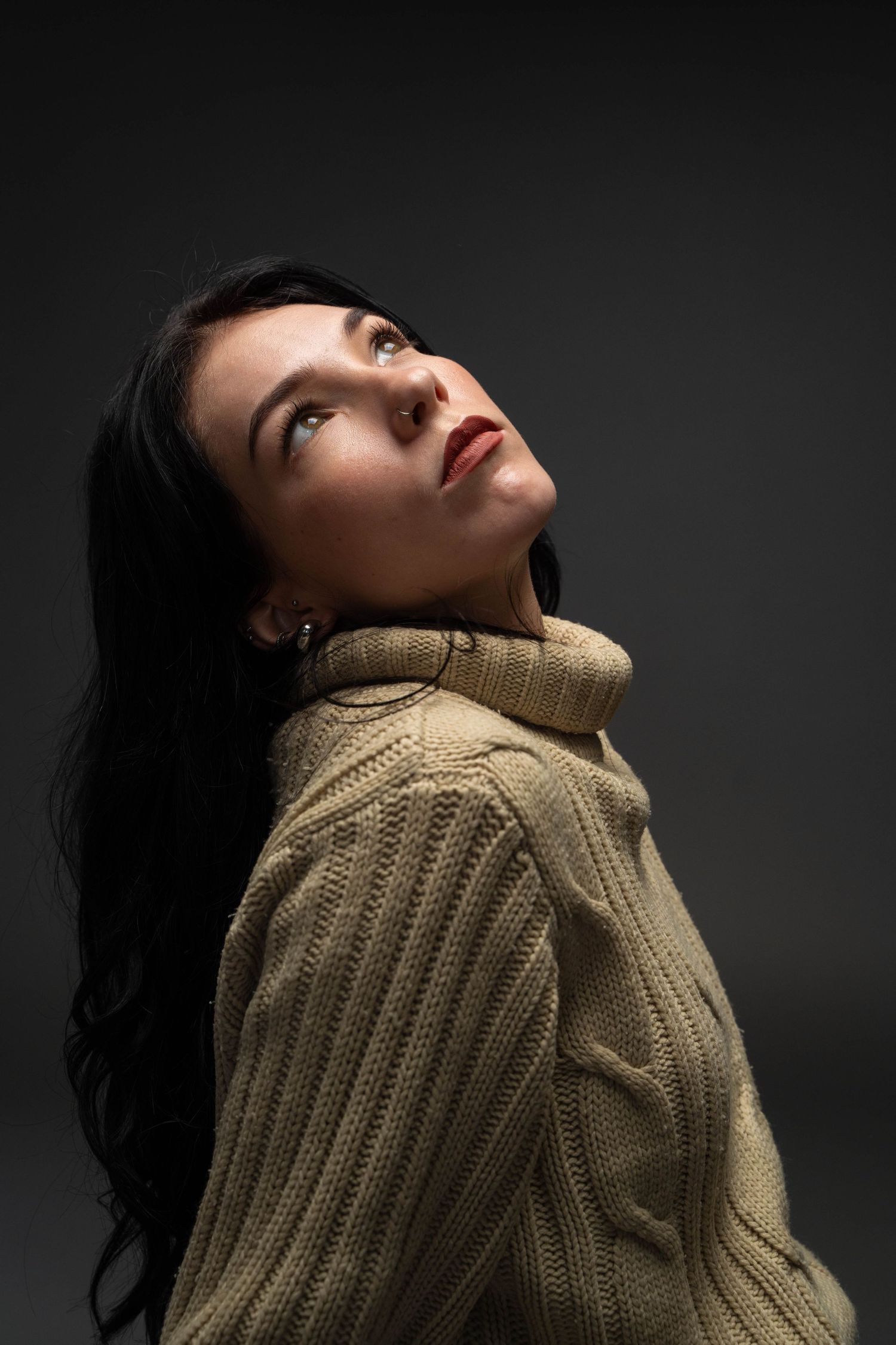 Artistic portrait in beige sweater with dramatic upward gaze against dark background.