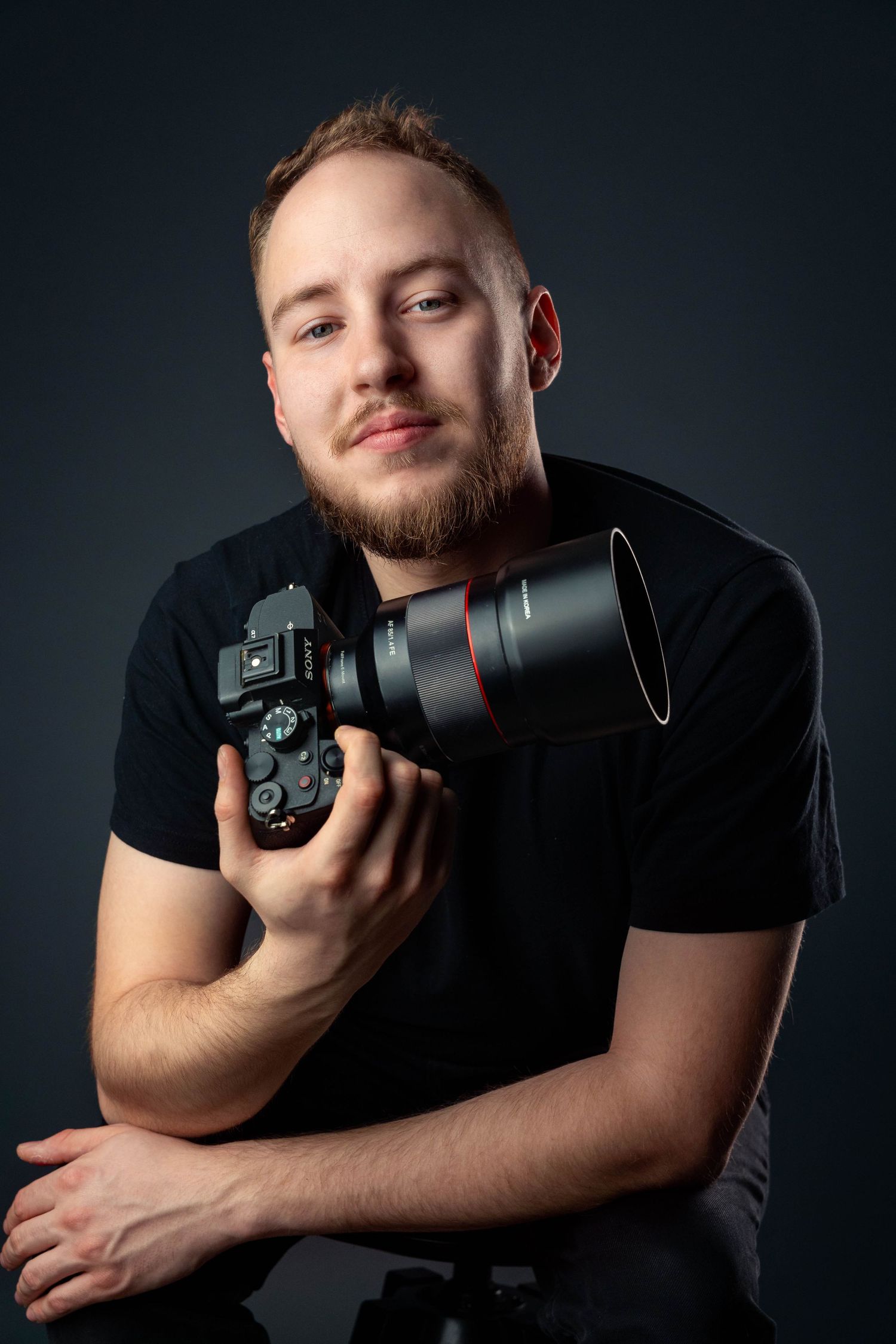 Professional photographer in black t-shirt holding DSLR camera with telephoto lens against dark studio background.