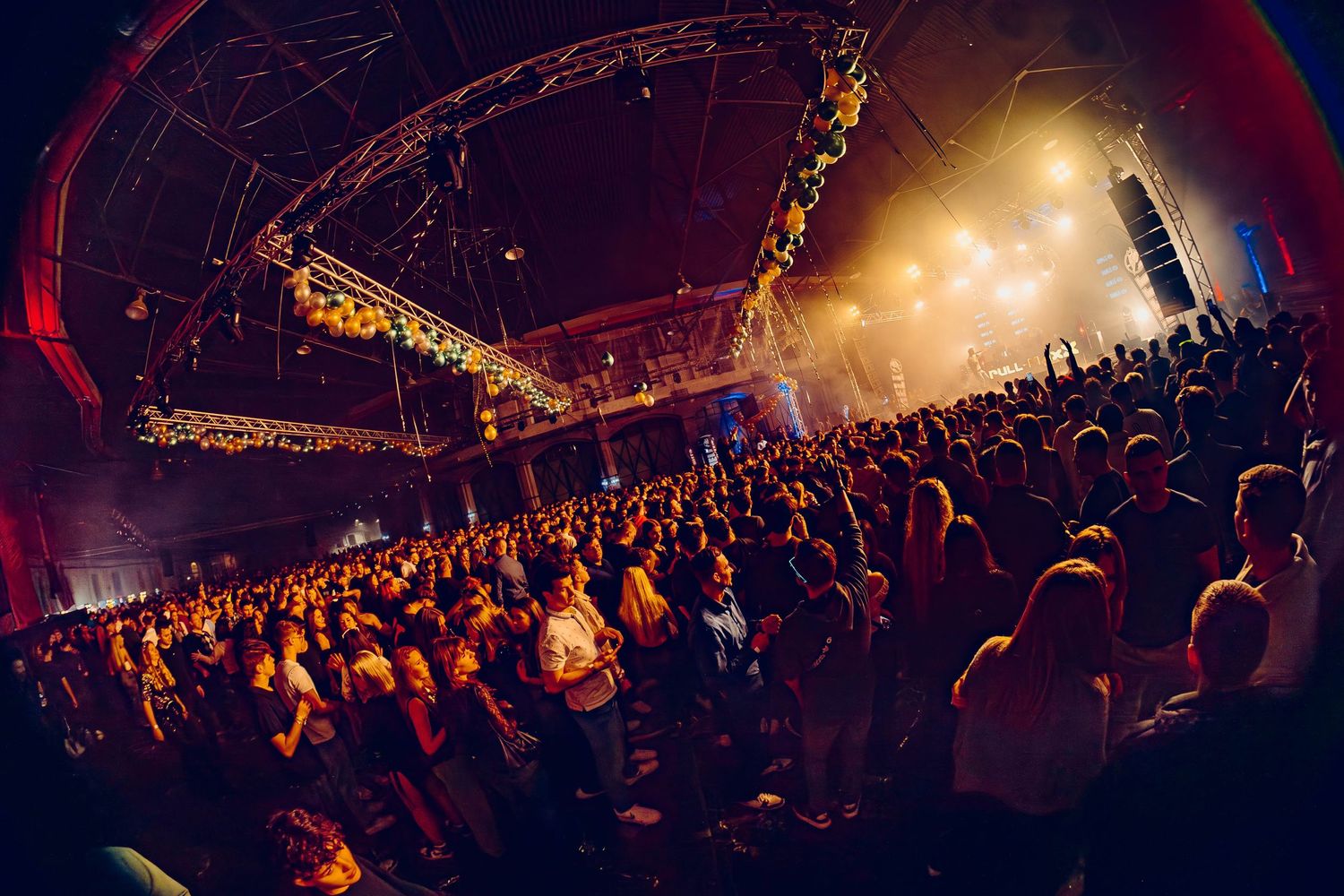 Fisheye view of a crowded concert venue with dramatic lighting and illuminated stage trusses overhead.