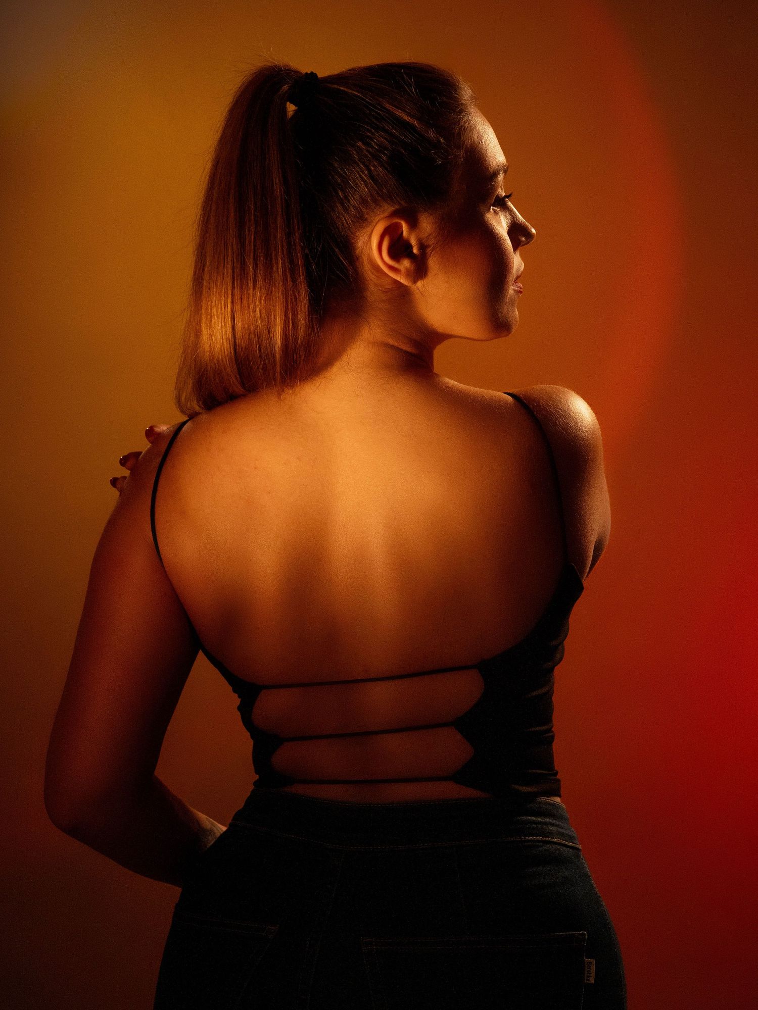 Back view portrait in warm amber lighting showing strappy dress detail against orange-red background.