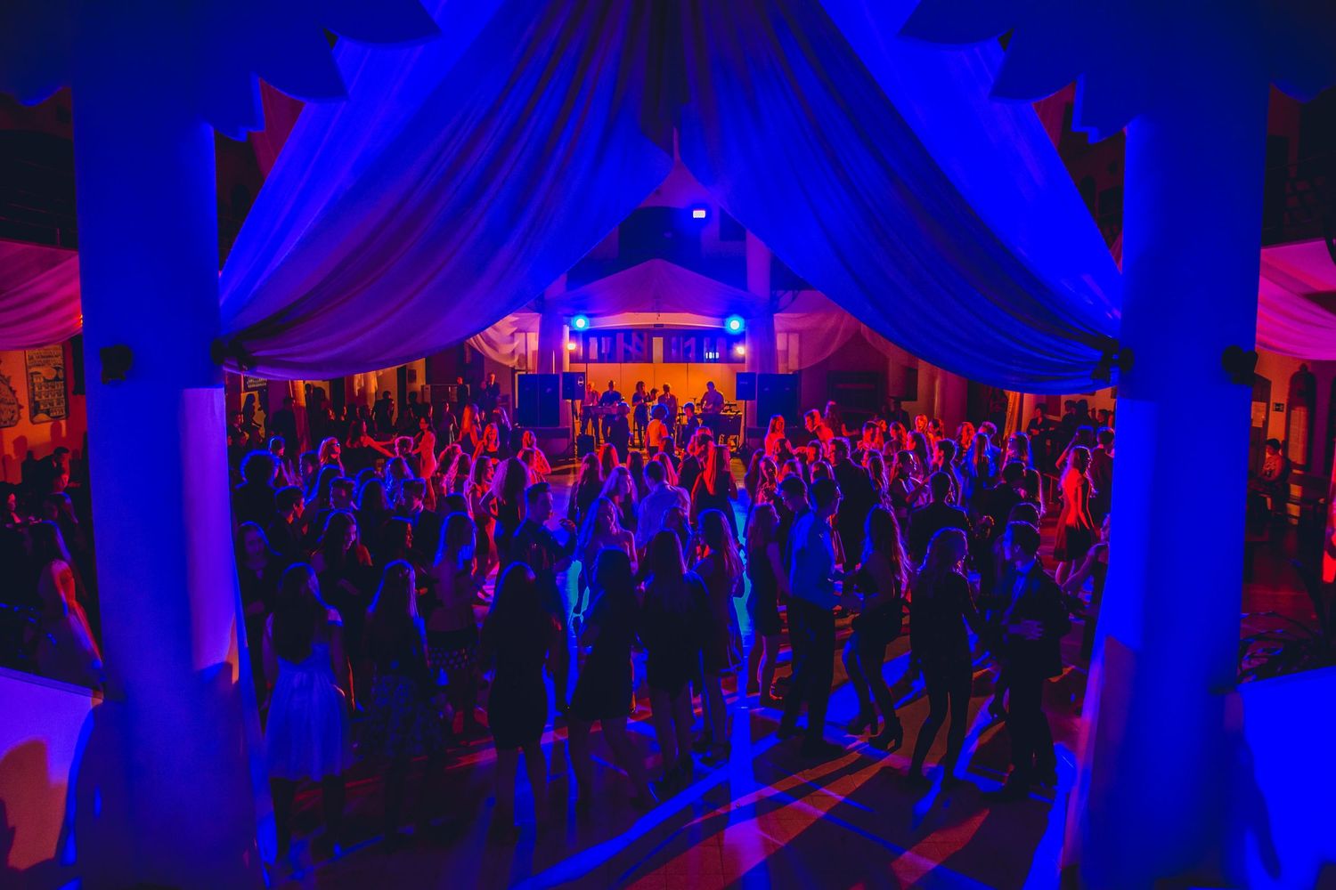 A crowded dance floor illuminated by vibrant blue and purple lighting under draped fabric at a nightclub event.
