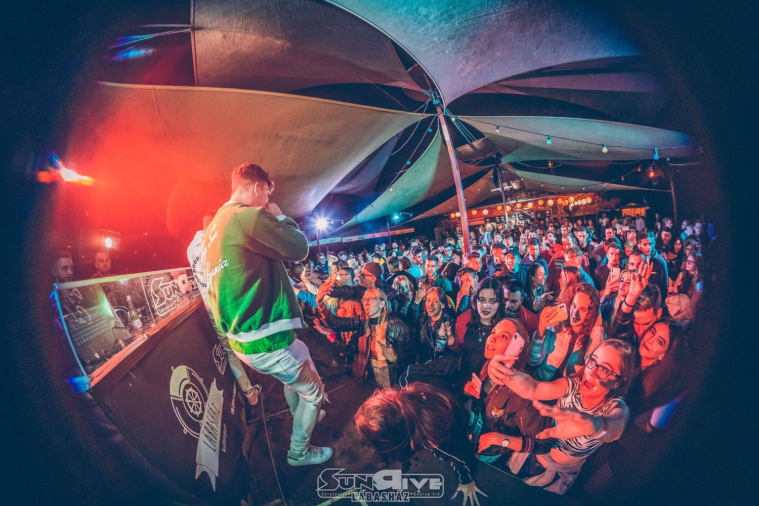 Vibrant concert scene with performer on stage and large crowd illuminated by red and blue lighting effects through fisheye lens.