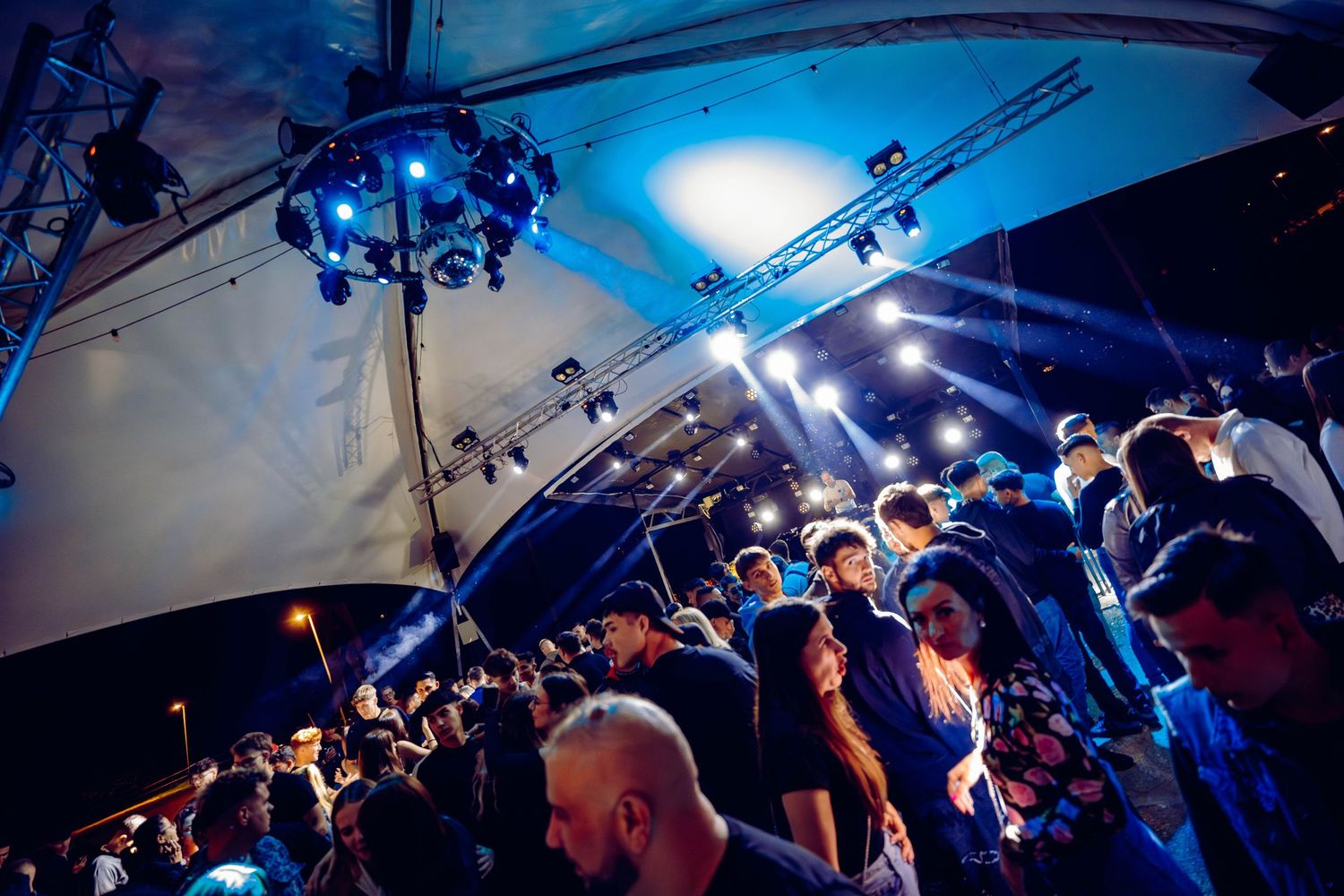 A lively crowd dances with blue lighting effects in a DJ performance at a nightclub venue.