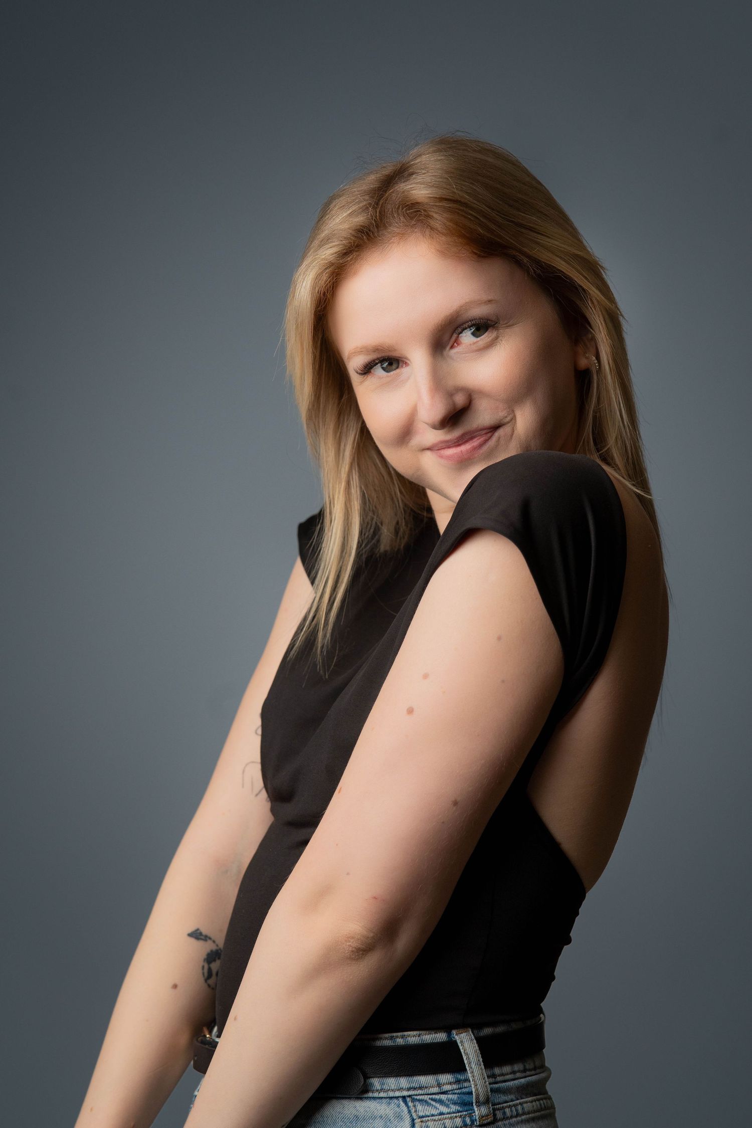 Portrait against gray backdrop showing person with blonde hair wearing black sleeveless top and jeans in casual pose.