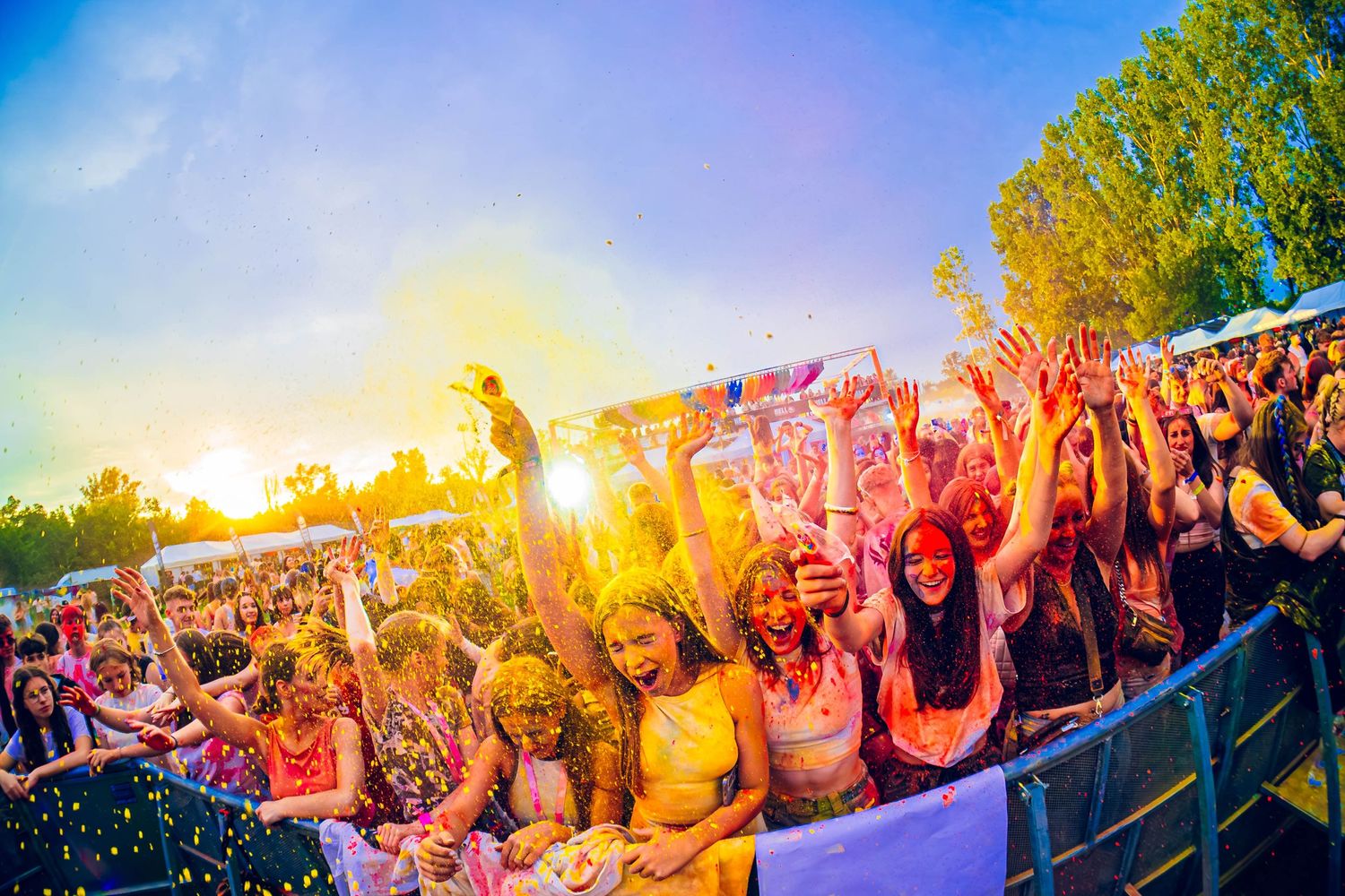 Festival crowd covered in colorful powder celebrating at sunset with vibrant sky.