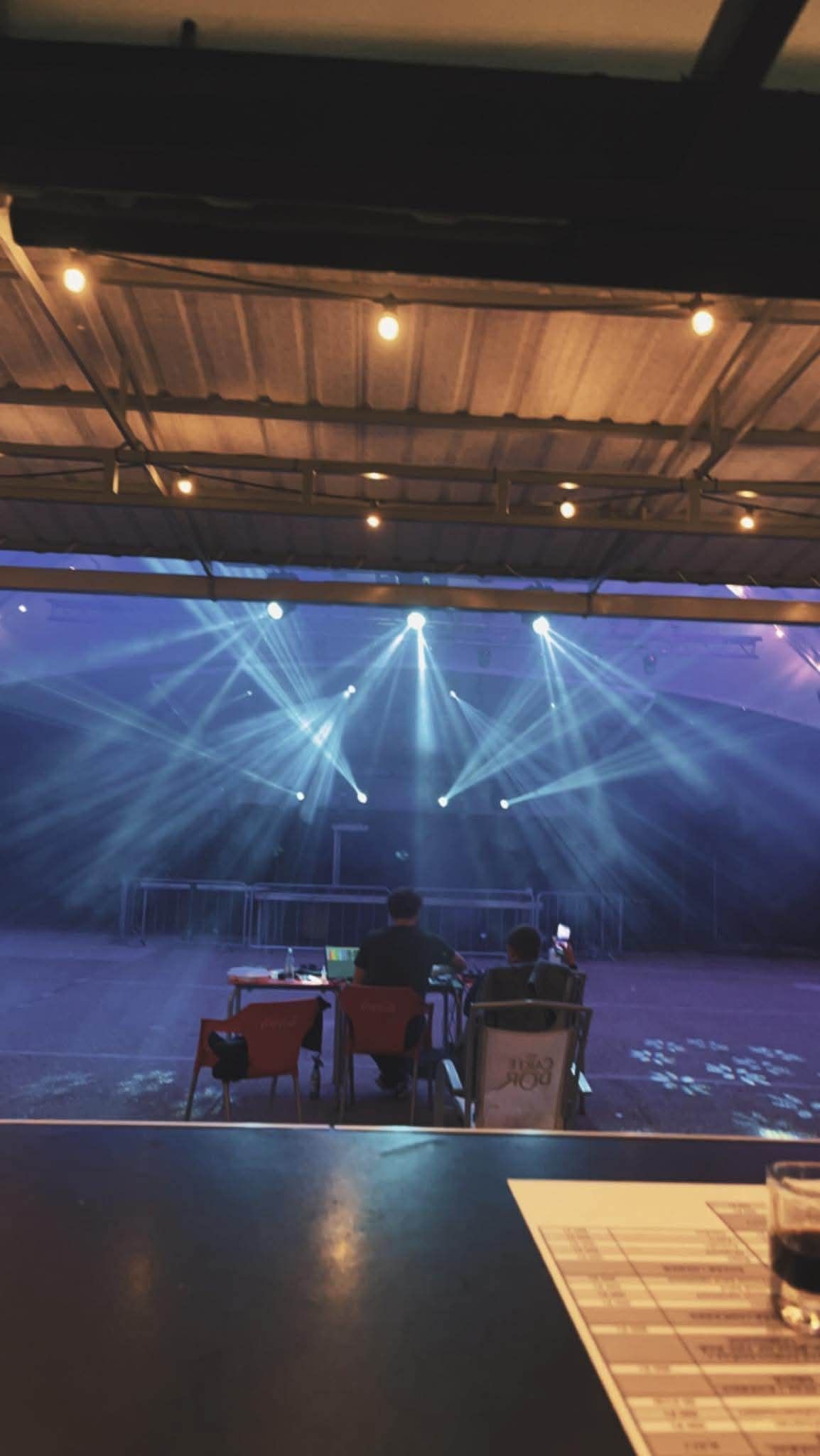 Blue spotlights shine dramatic beams across an empty stage as viewed from behind a production desk or control area.