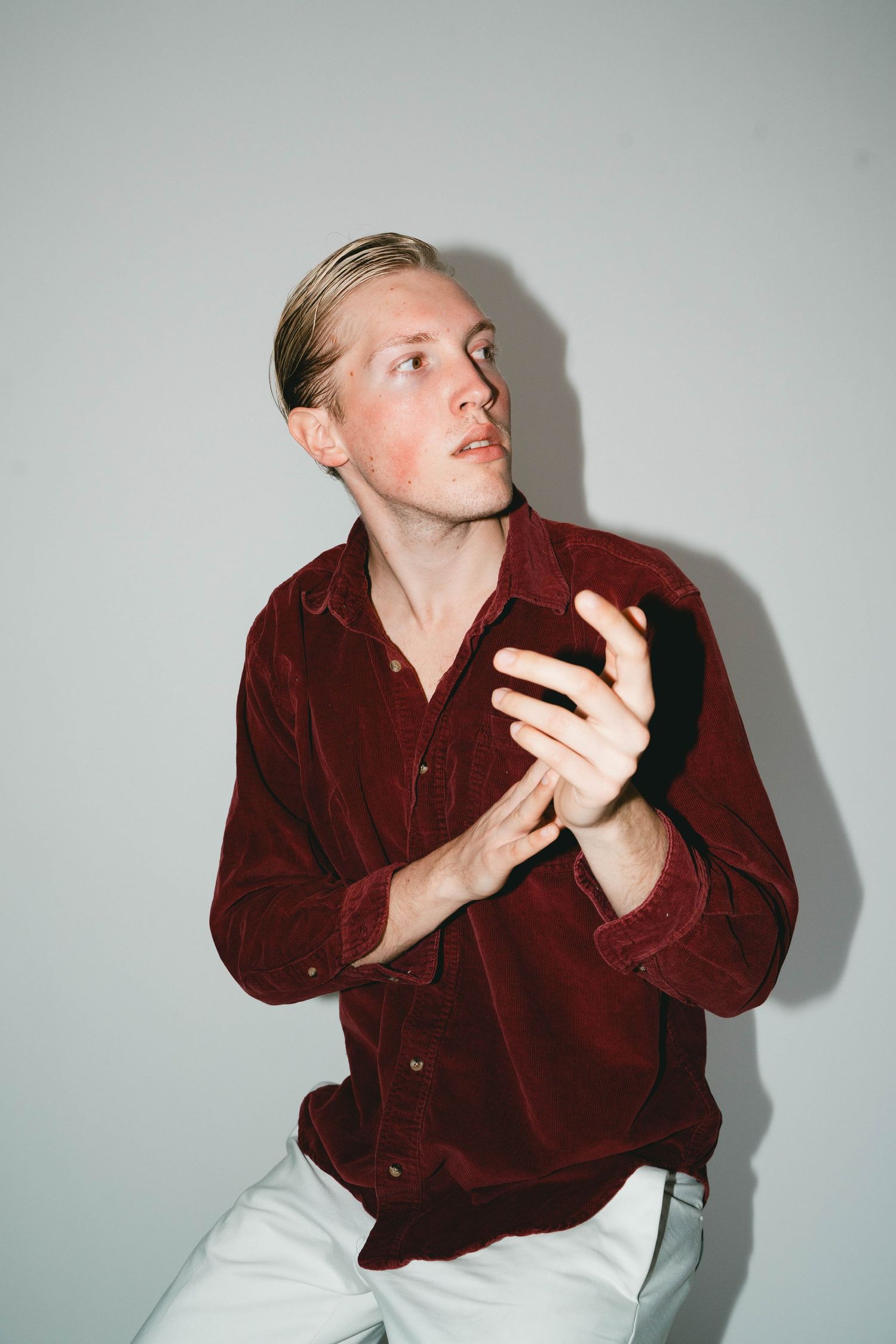 A person wearing a burgundy corduroy button-up shirt and white pants poses expressively against a plain wall.