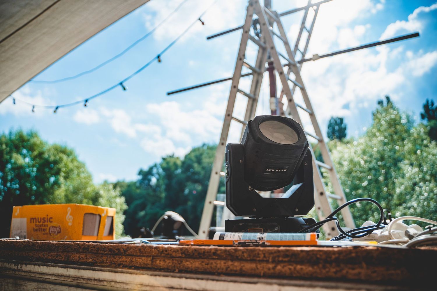 Professional stage lighting equipment and sound gear set up outdoors with string lights and trees in background.