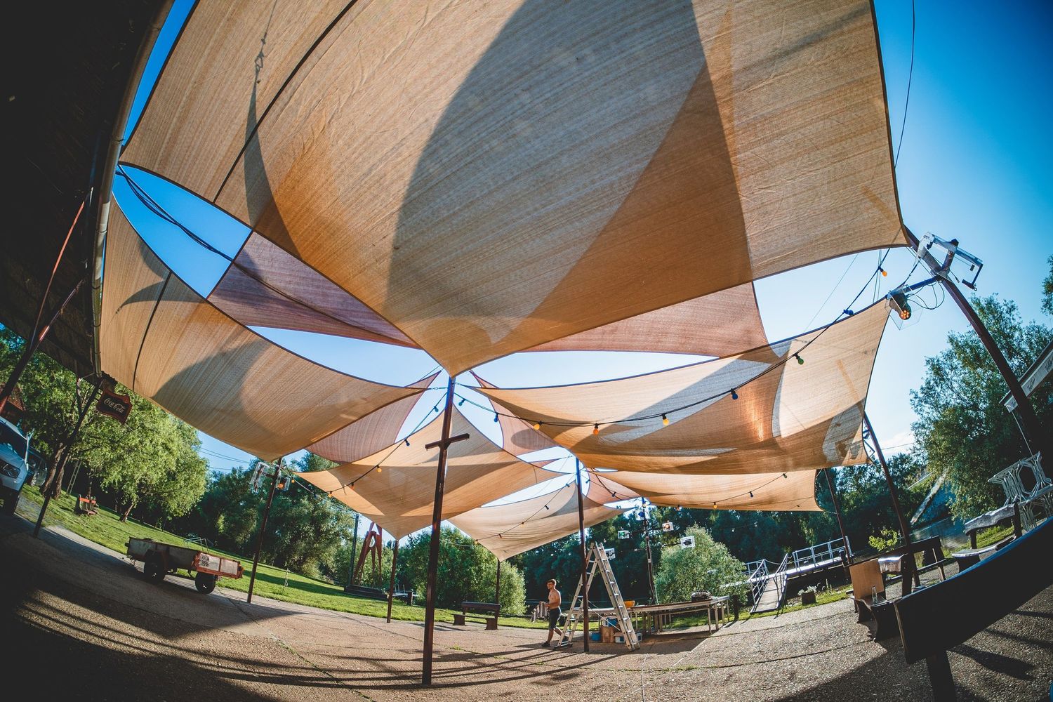 Curved fabric shade sails provide overhead coverage in an outdoor park area under bright blue sky.